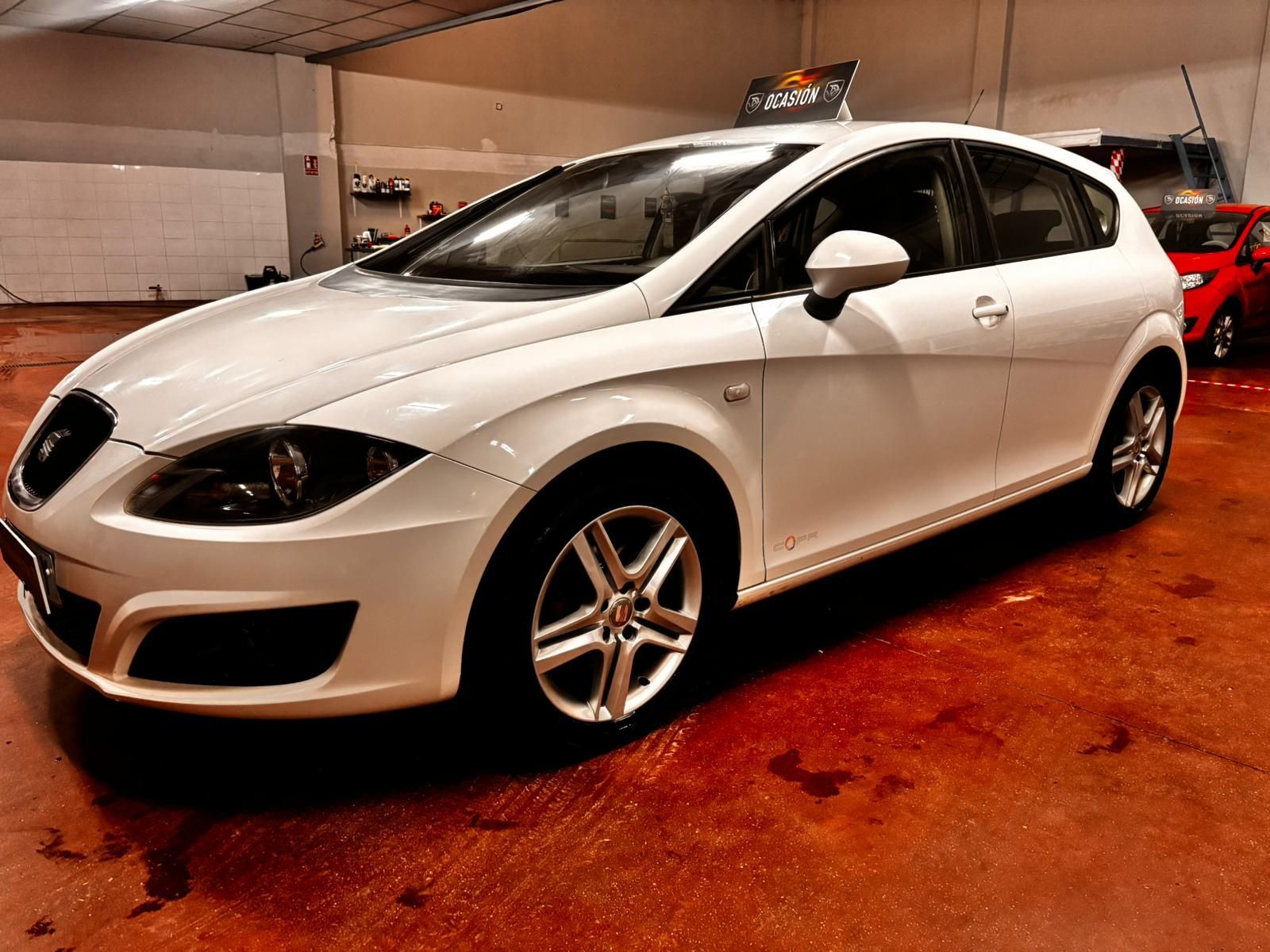 Un Seat León hatchback blanco estacionado en el interior sobre un suelo rojizo, visto desde un ángulo de tres cuartos frontal.