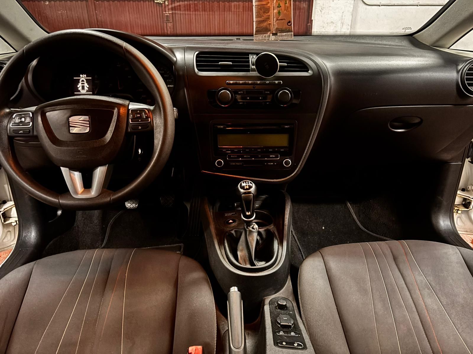 Vista interior del habitáculo de un Seat de tonos oscuros, con volante, palanca de cambios manual y pantalla en el salpicadero.