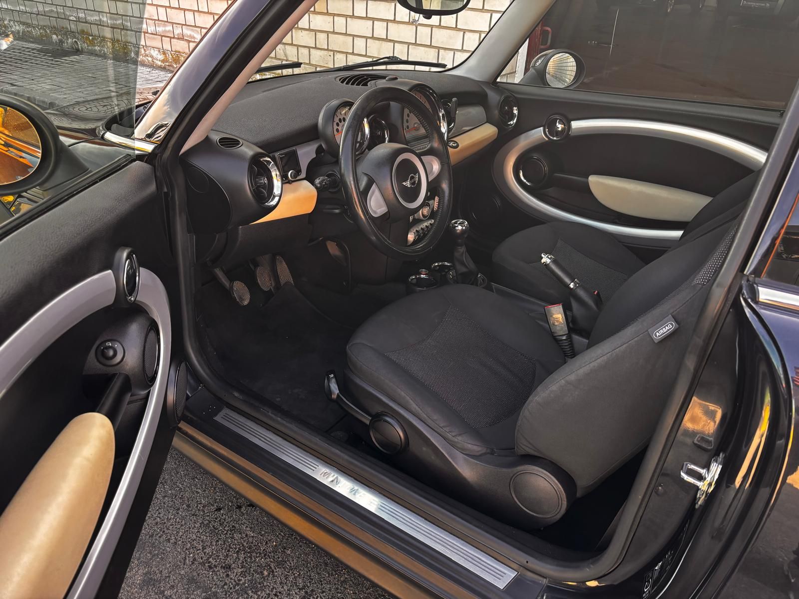 Interior de un Mini Cooper con asientos deportivos de cuero negro, detalles en color beige y un velocímetro circular prominente montado en el centro.