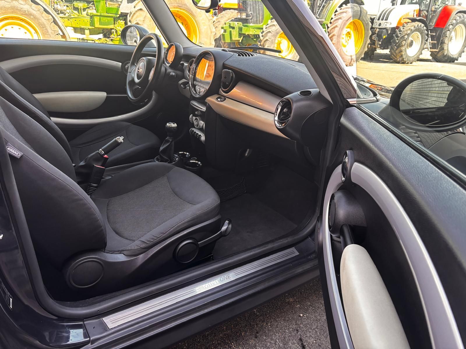 El interior de un Mini Cooper negro con asientos individuales de tela oscura estampada, palanca de cambios manual y salpicadero redondeado.