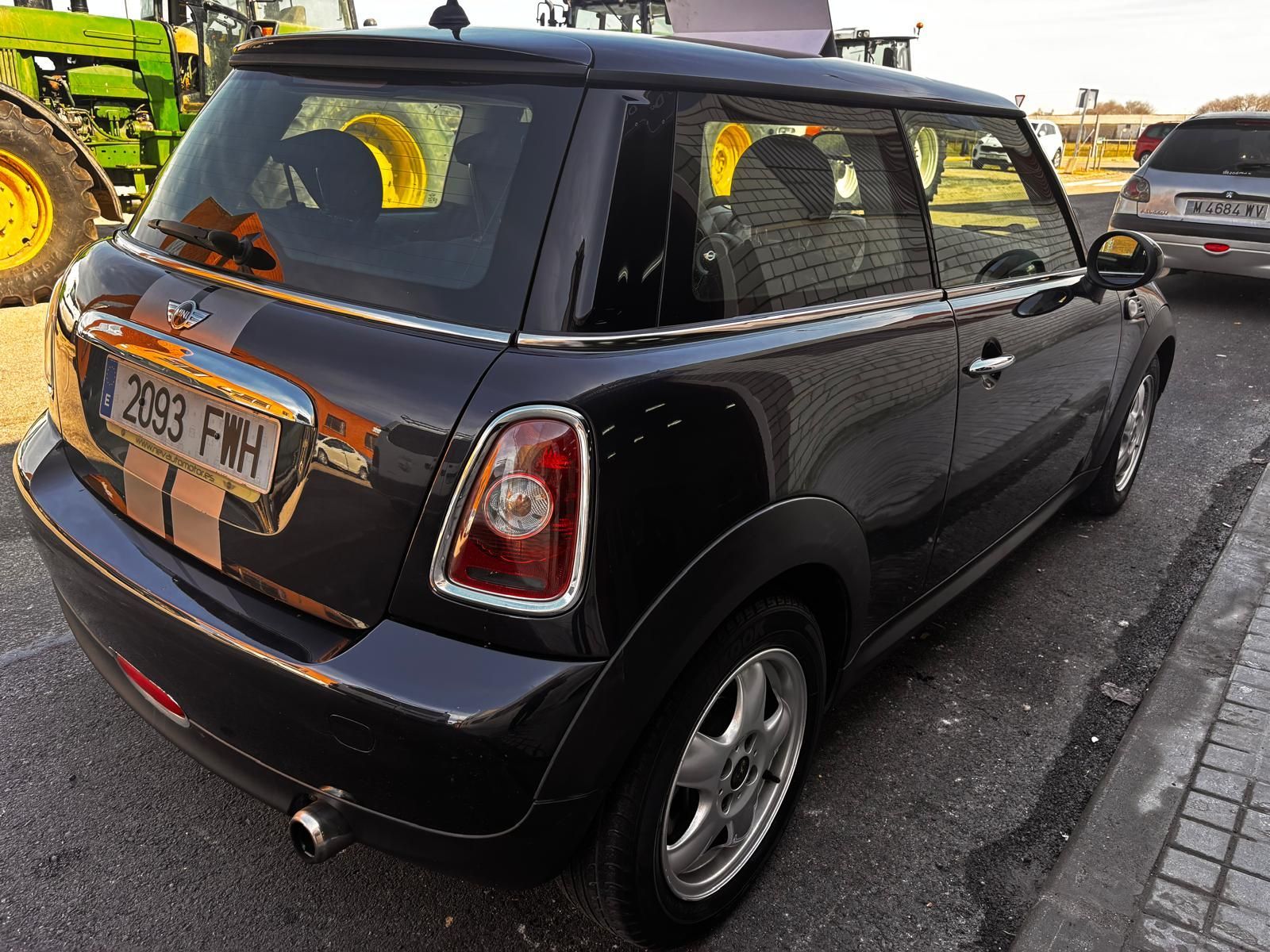 Un Mini Cooper negro estacionado al aire libre, visto desde una perspectiva trasera de tres cuartos, con la rueda de un tractor visible al fondo.