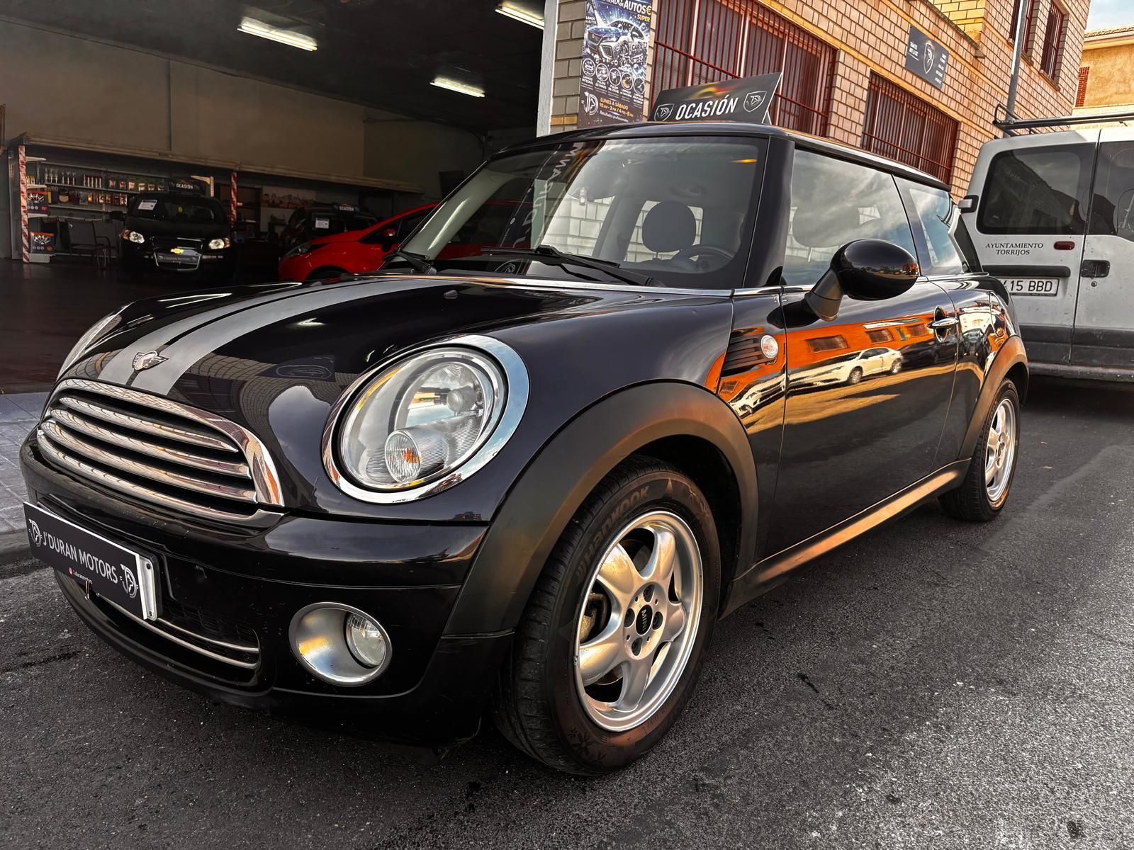 Un Mini Cooper negro con franjas de carreras blancas aparcado frente a un edificio junto a una furgoneta blanca.
