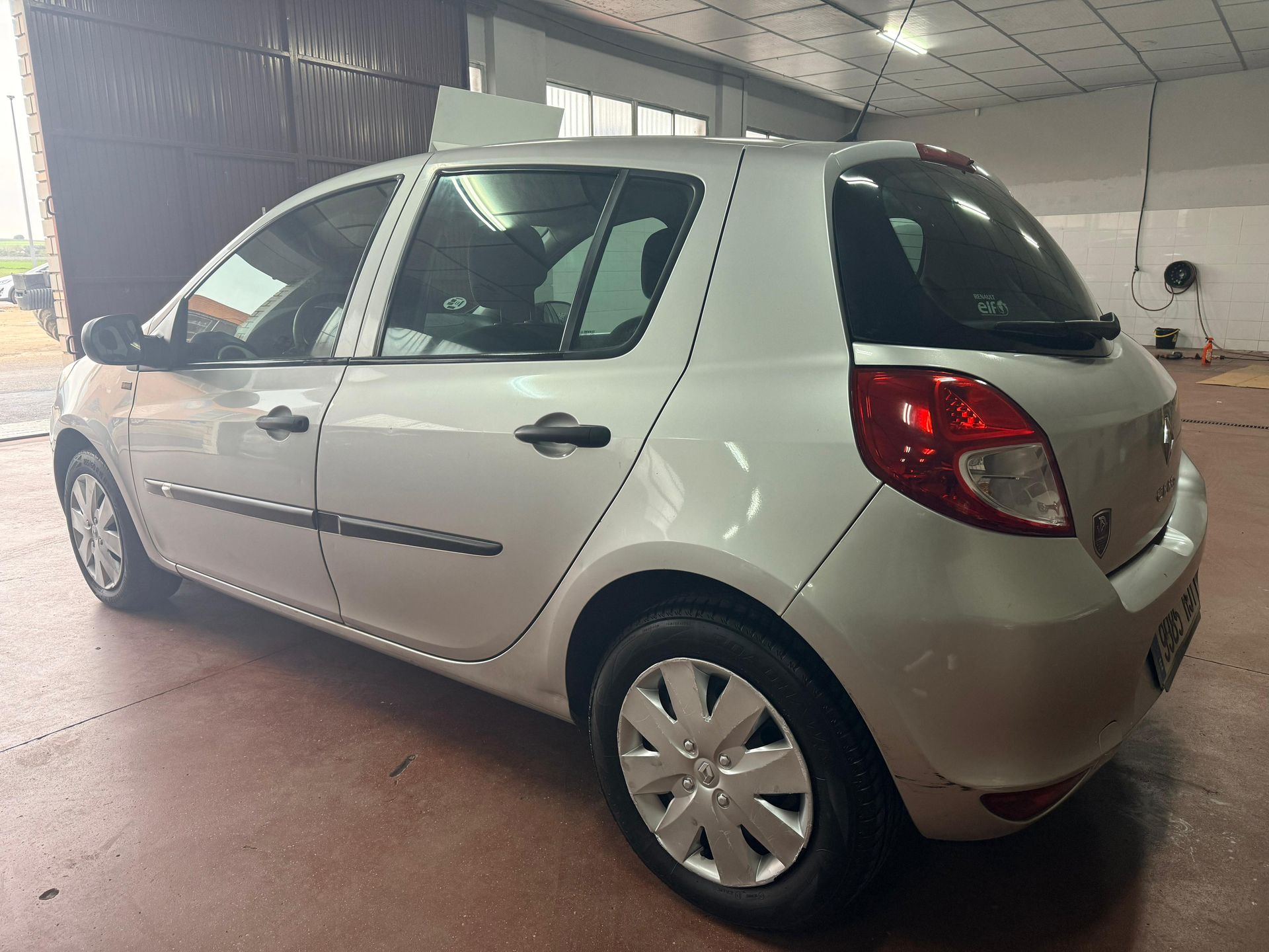 Un Renault Clio hatchback plateado estacionado ligeramente inclinado dentro de un garaje o taller bien iluminado.