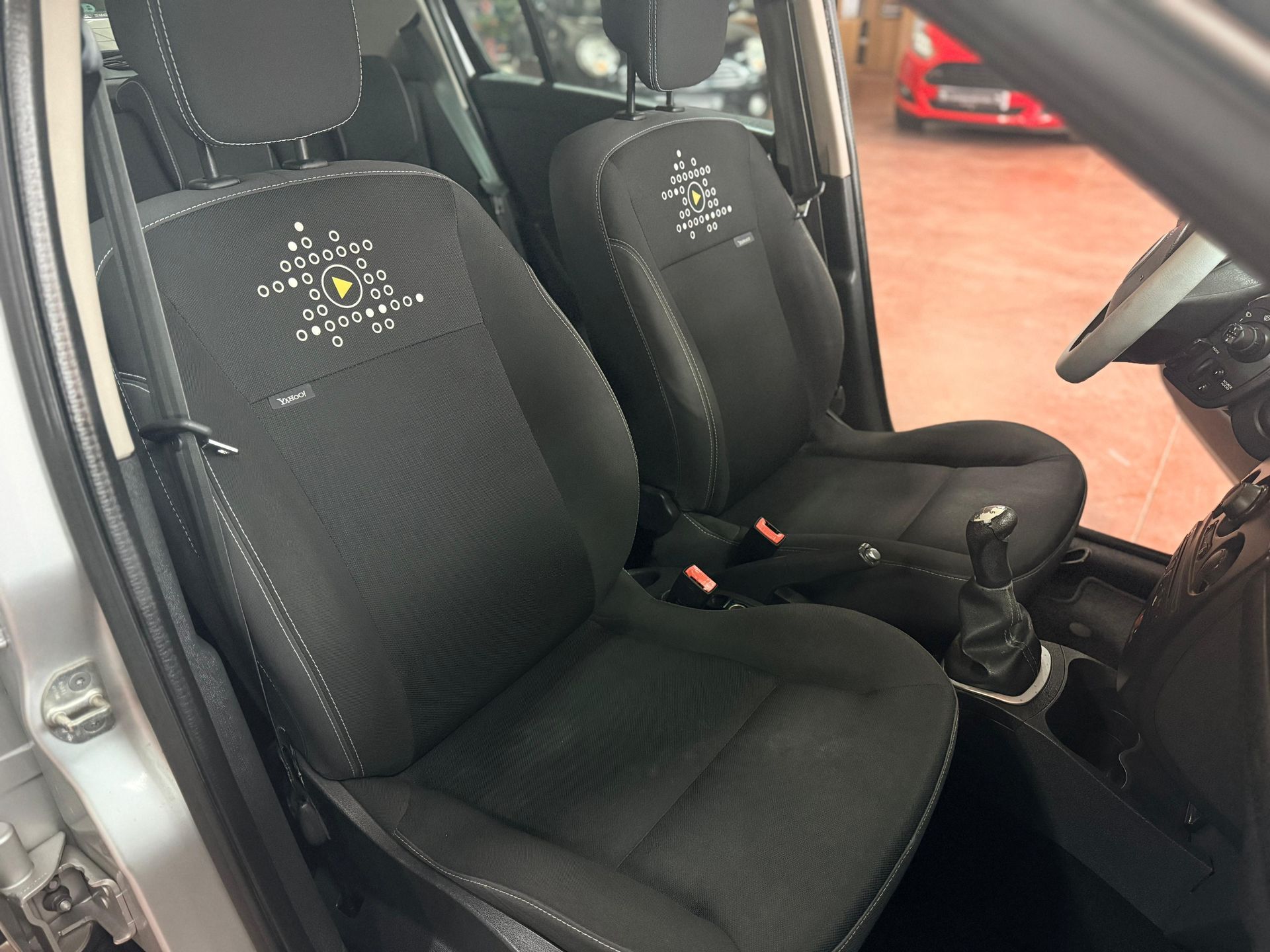 El interior del coche muestra dos asientos delanteros negros tapizados en tela con motivos geométricos blancos y una palanca de cambios manual.