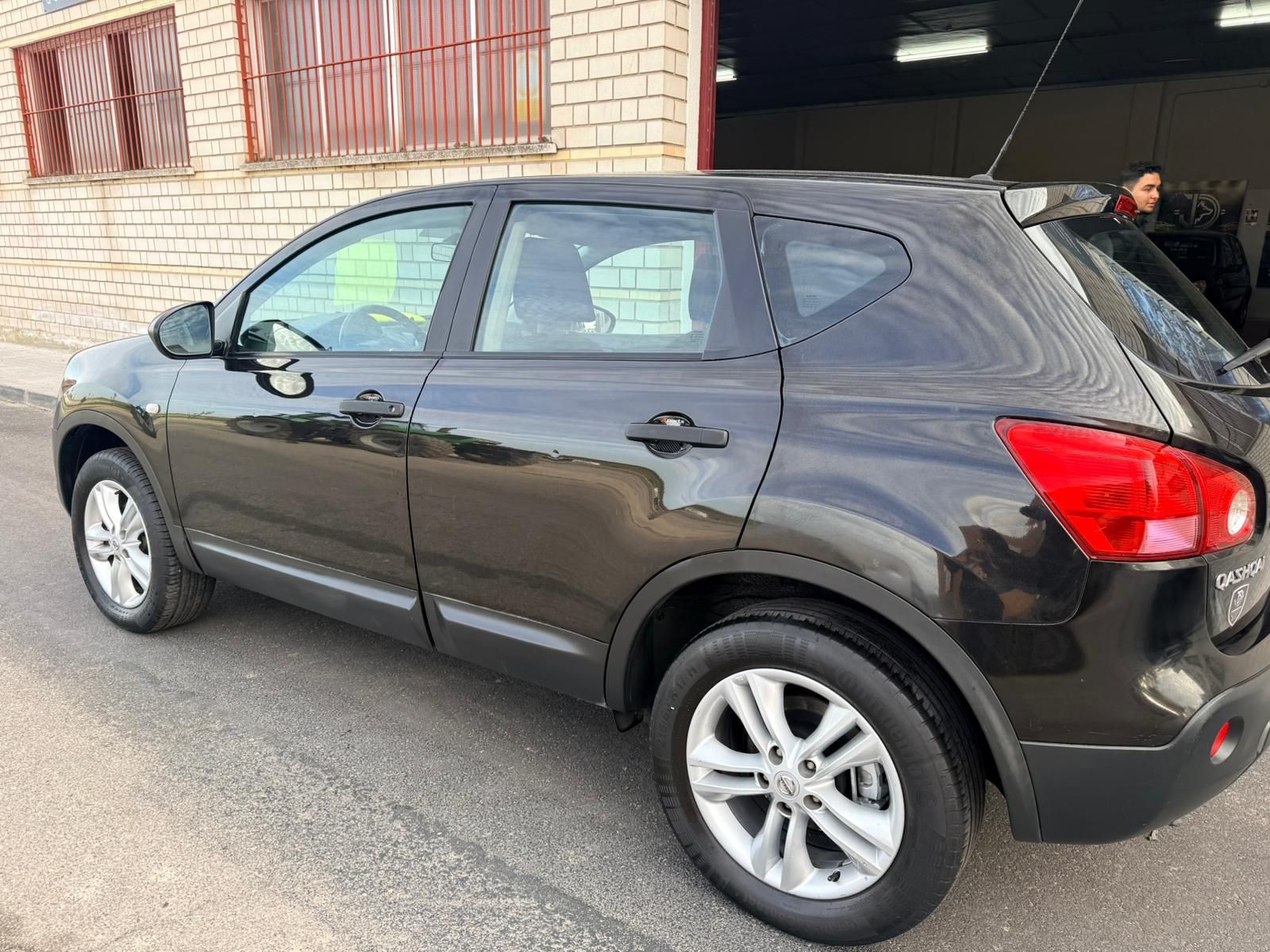 Un SUV Nissan Qashqai negro estacionado sobre una superficie de asfalto frente a un edificio de ladrillo.