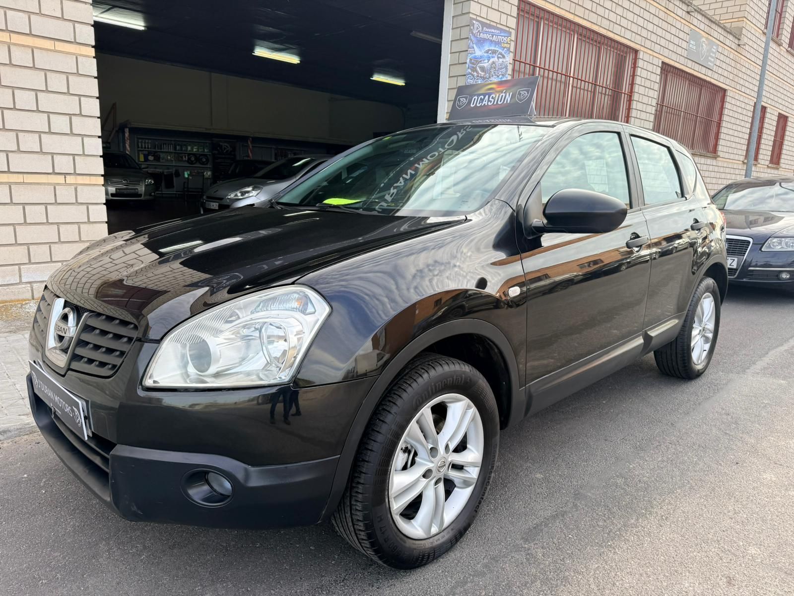 Un SUV crossover Nissan Qashqai negro estacionado frente a un edificio de ladrillo en un día soleado.