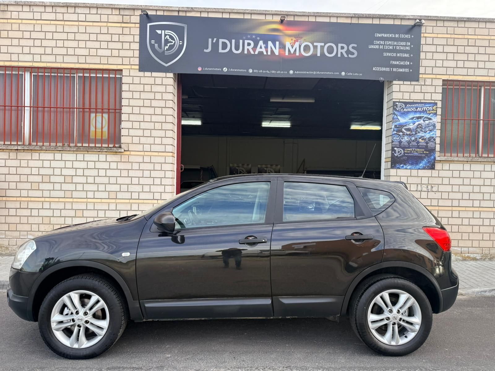 Un SUV gris oscuro estacionado frente a un edificio de ladrillo con un letrero de J'Duran Motors sobre la entrada del garaje.