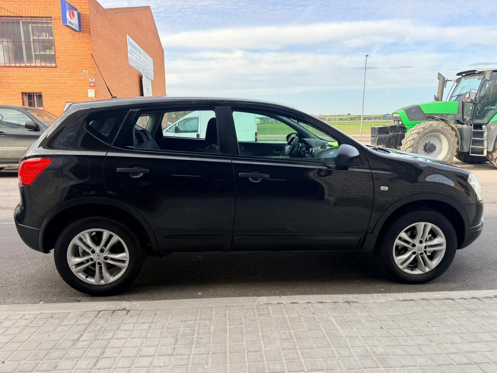 Un SUV negro estacionado en una calle pavimentada cerca de un edificio de ladrillos, con un tractor verde visible al fondo.