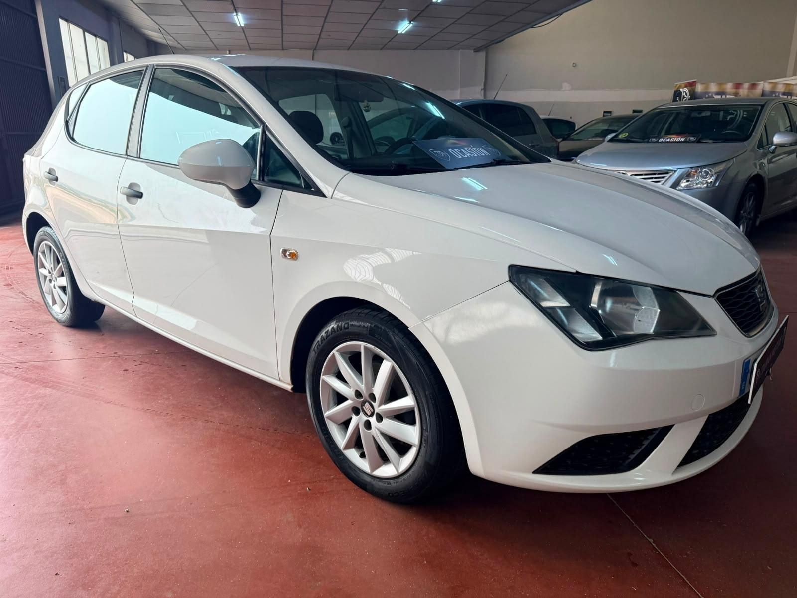 Un Renault Clio hatchback plateado estacionado dentro de una sala de exposición de automóviles, con un coche rojo y un coche blanco visibles al fondo.