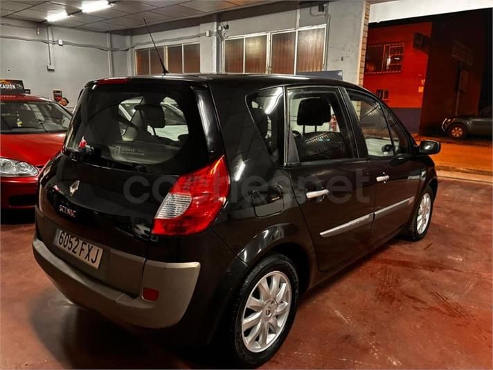 Minivan Renault Scénic negra estacionada en interior con parachoques trasero inferior beige, al lado de un coche rojo.