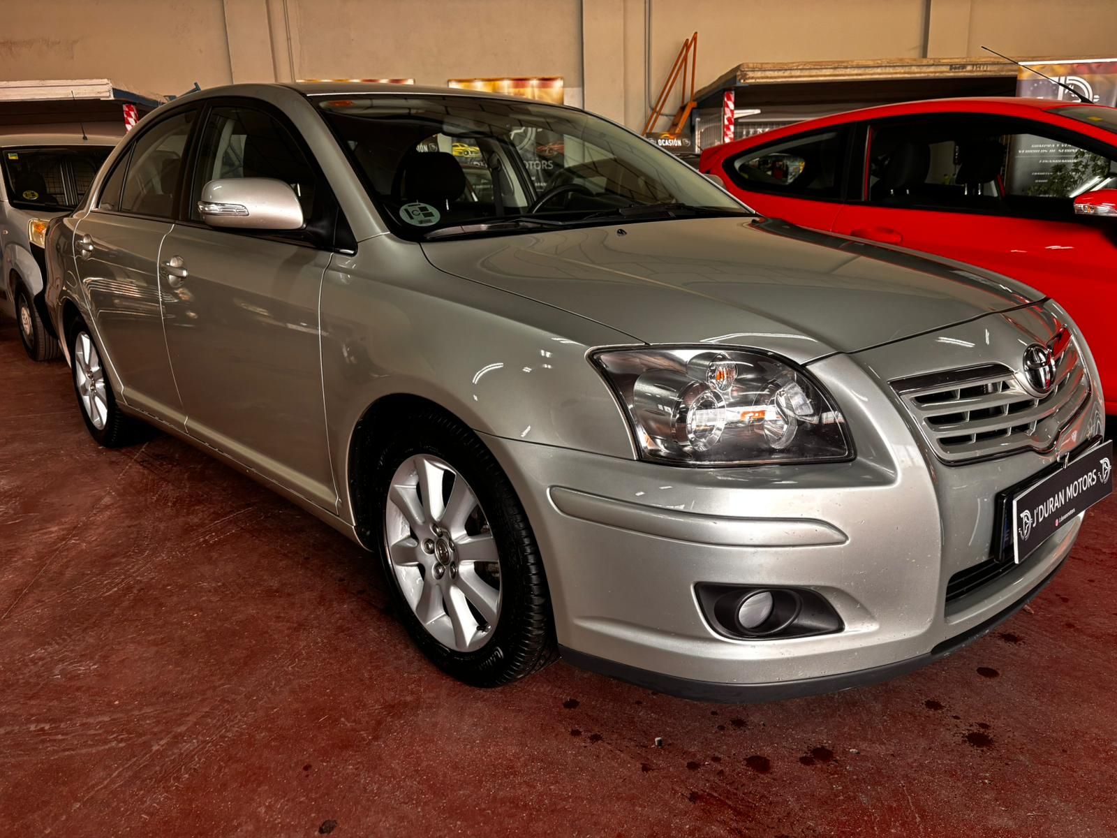 Un sedán Toyota Avensis plateado estacionado en el interior, sobre un suelo rojo oscuro, orientado hacia el espectador.