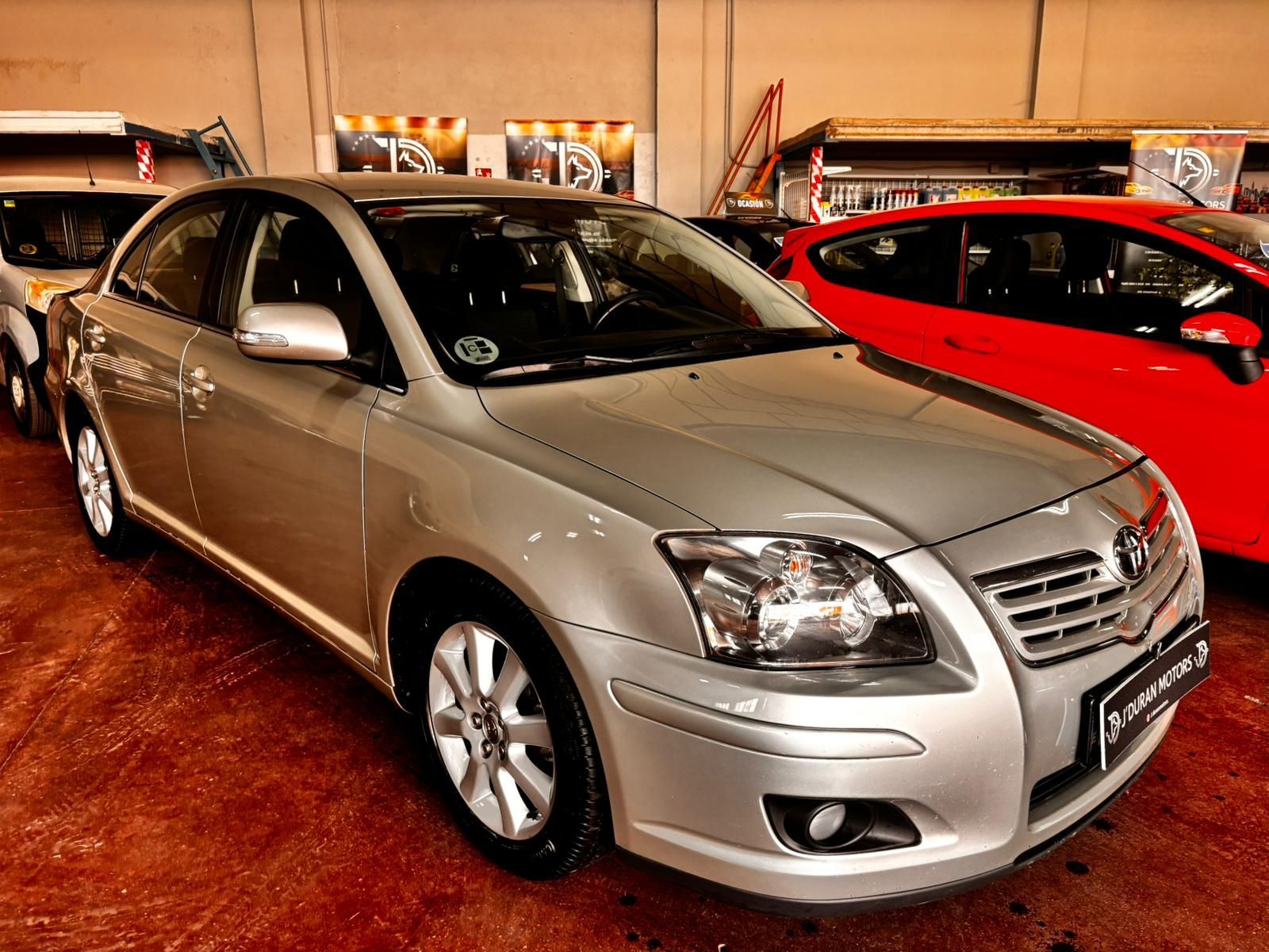 Un Toyota Avensis plateado estacionado en el interior sobre un suelo rojo, con un coche rojo visible al lado.
