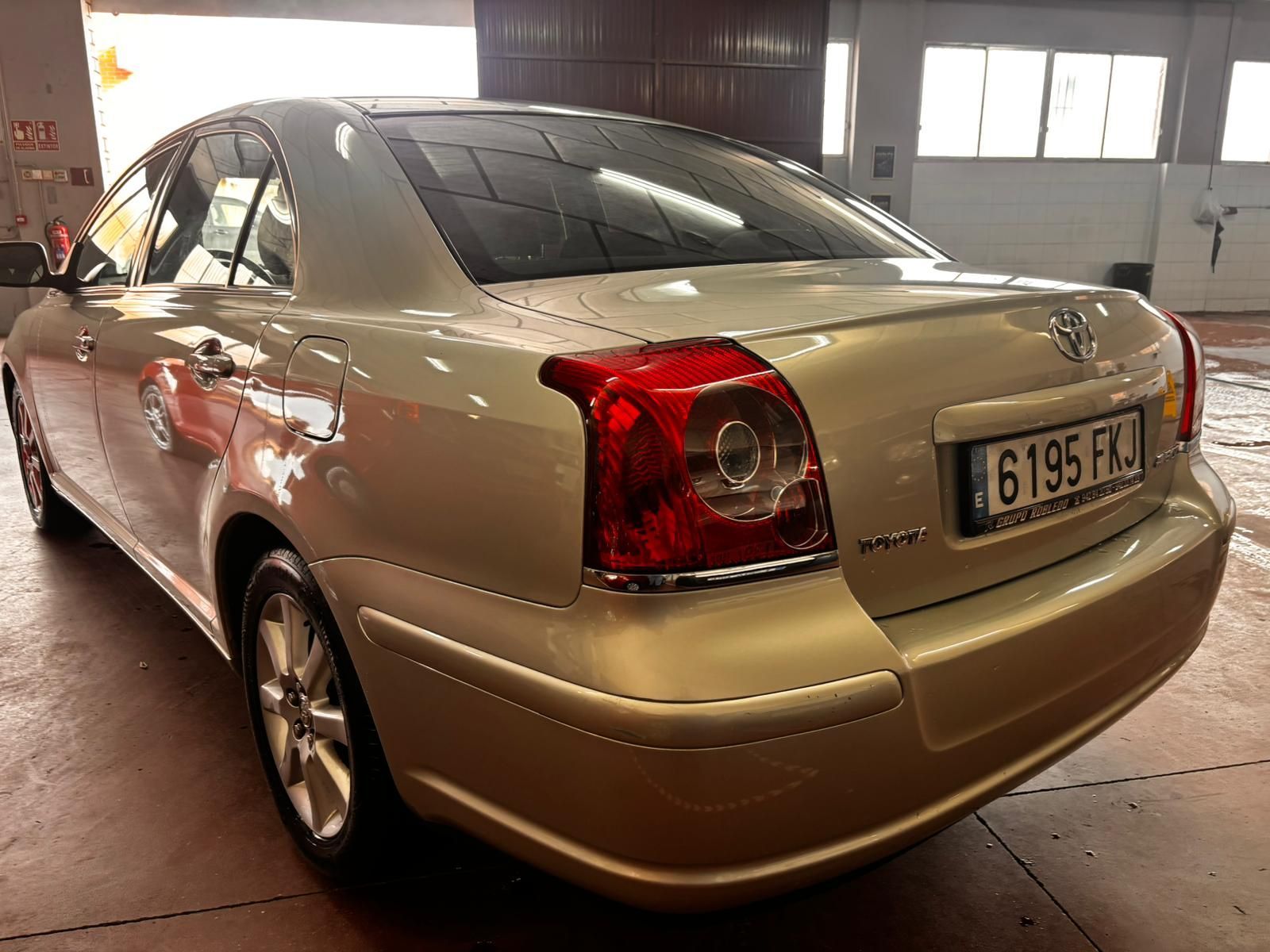 Vista trasera de tres cuartos de un sedán Toyota Avensis dorado estacionado en el interior sobre un suelo de hormigón.