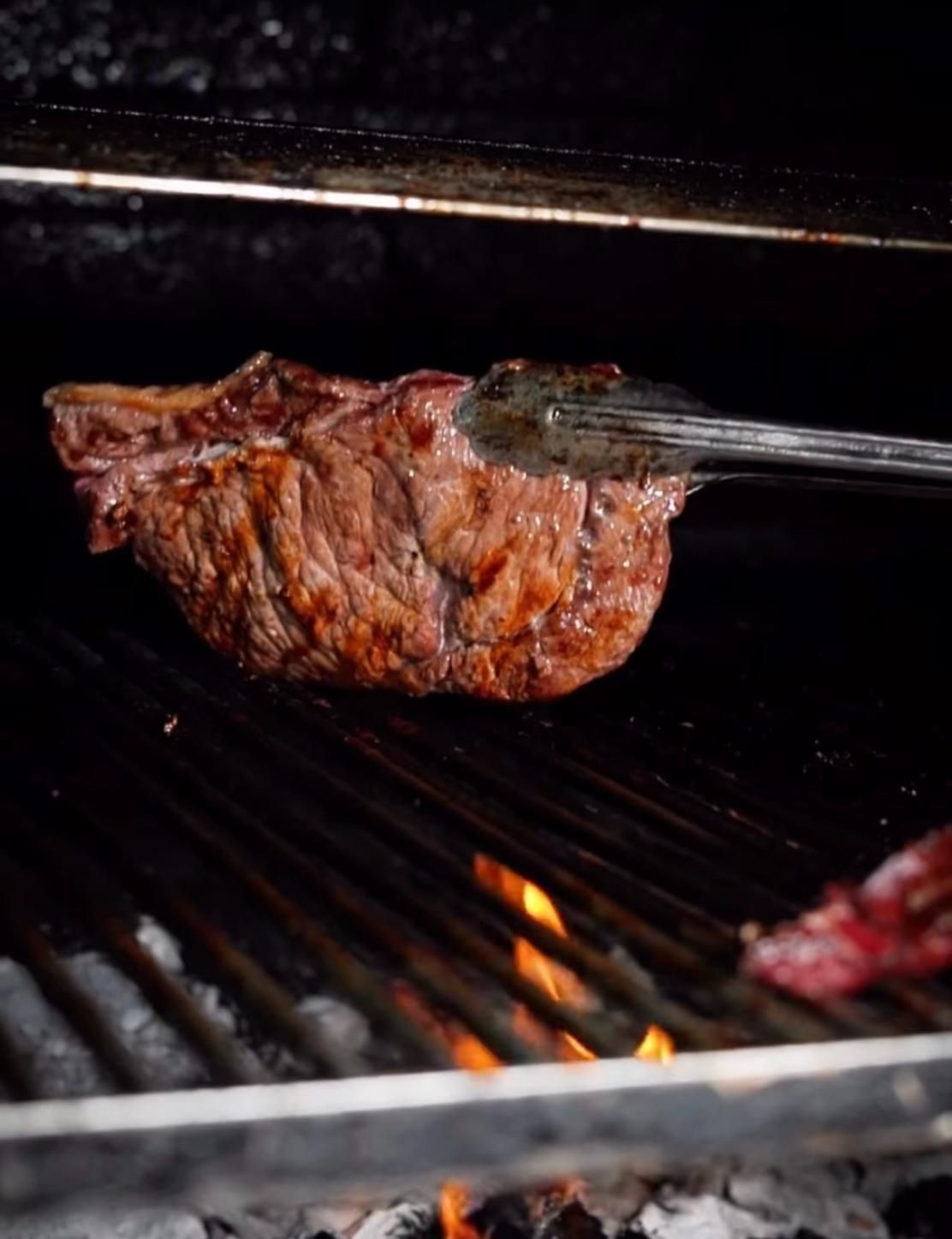 Filete sellándose en la parrilla, sujeto con pinzas metálicas sobre brasas incandescentes.