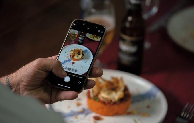 Una mano sostiene un teléfono inteligente fotografiando un postre emplatado sobre una mesa.