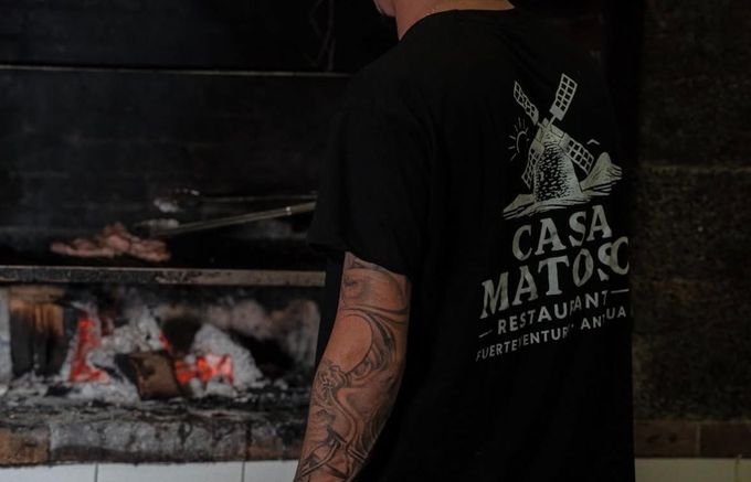 Persona asando carne en una barbacoa de carbón, vistiendo una camisa negra de Casa Matos.