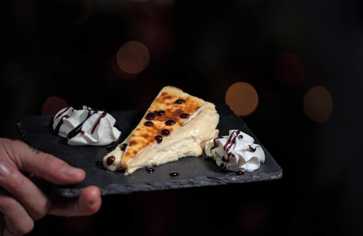 Mano sosteniendo una pizarra con porciones de tarta de queso y crema batida sobre un fondo oscuro.