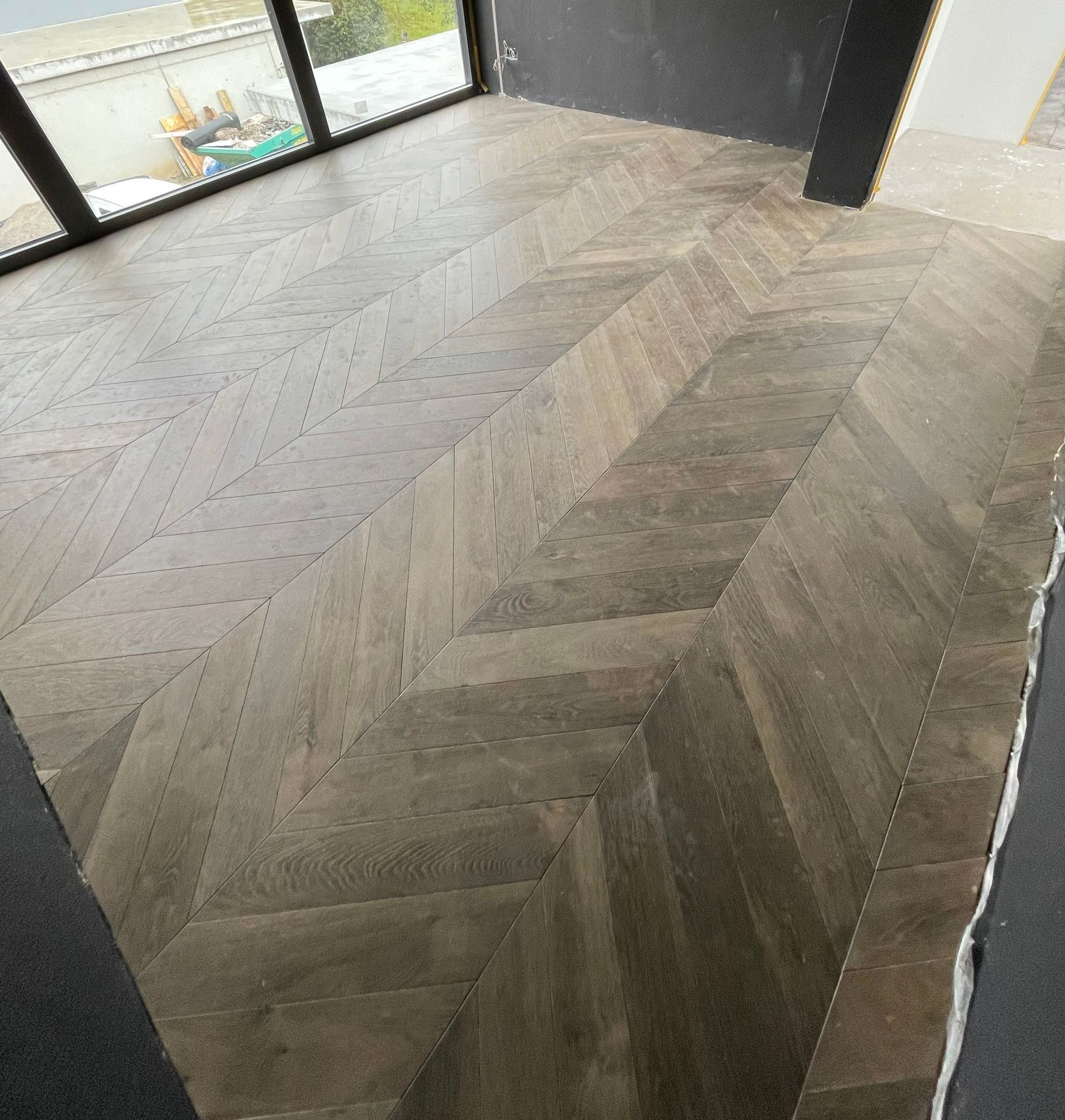 Ein Holzboden mit Fischgrätmuster in einem Raum mit großem Fenster.