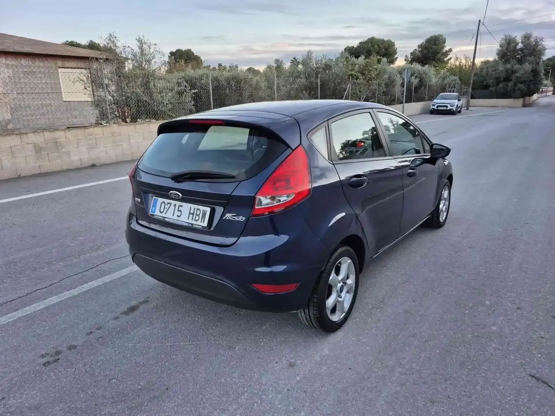 Un Ford Fiesta azul oscuro estacionado en una carretera asfaltada; al fondo hay árboles y un edificio.