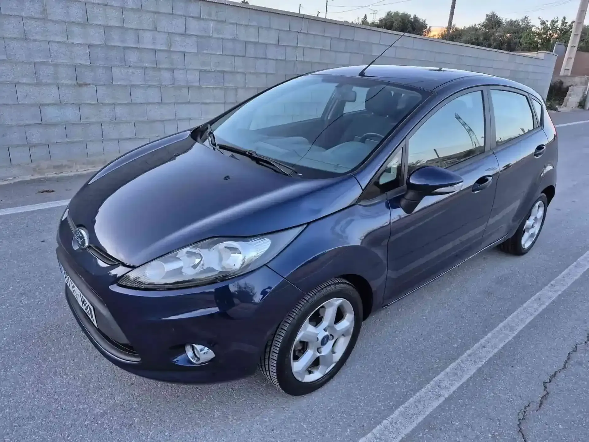 Ford Fiesta azul estacionado en el asfalto cerca de una pared de ladrillos.