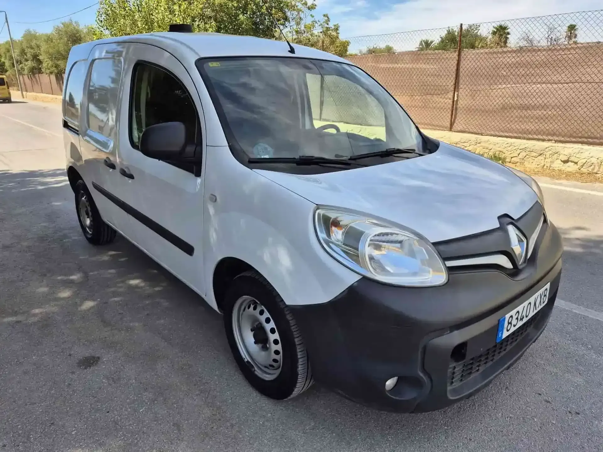 Furgoneta Renault Kangoo blanca estacionada en una carretera asfaltada. Parachoques y molduras negros. Exterior soleado.