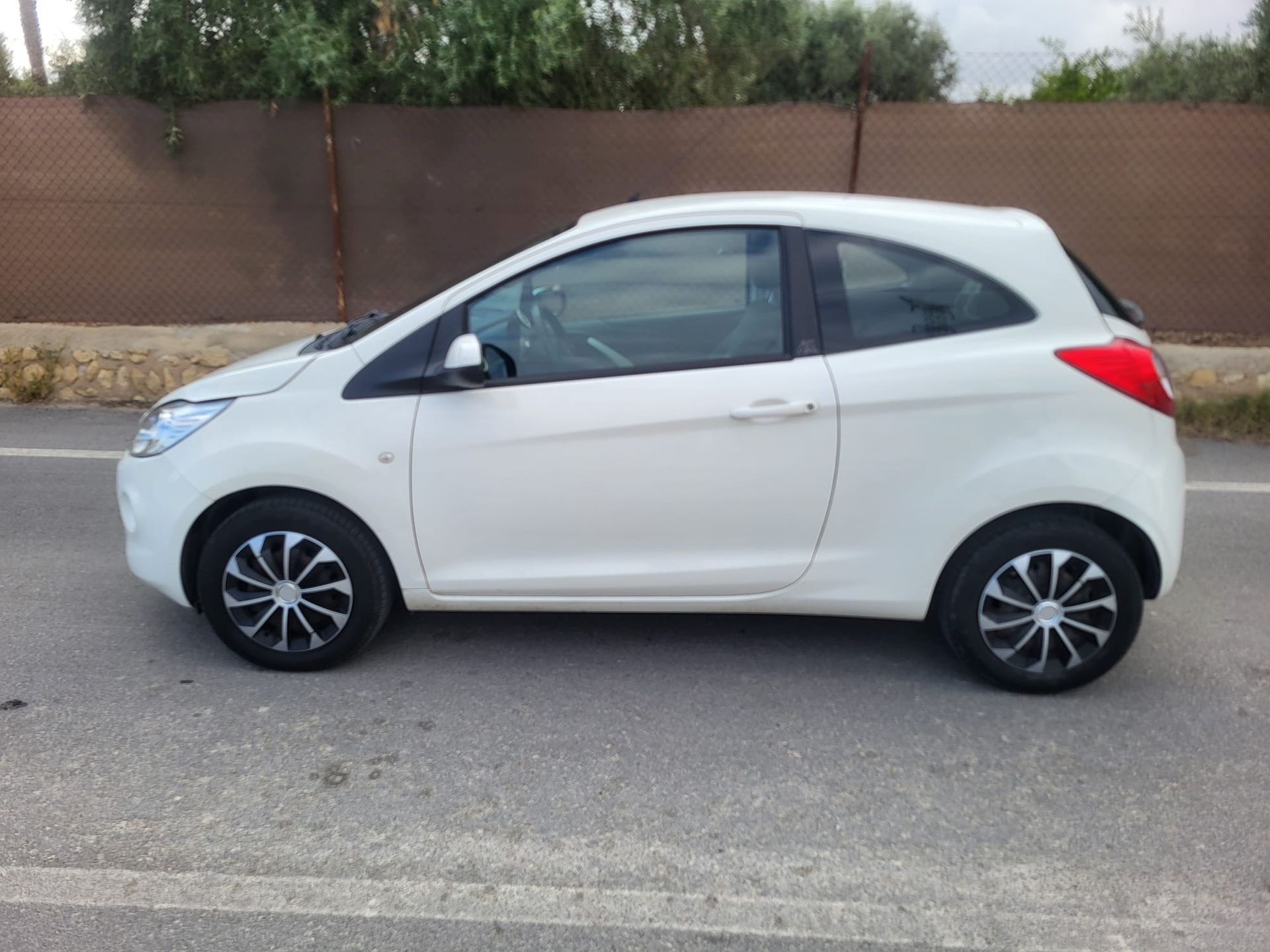 Un Ford Ka blanco de dos puertas estacionado sobre el asfalto, contra un fondo borroso de vegetación y una valla.