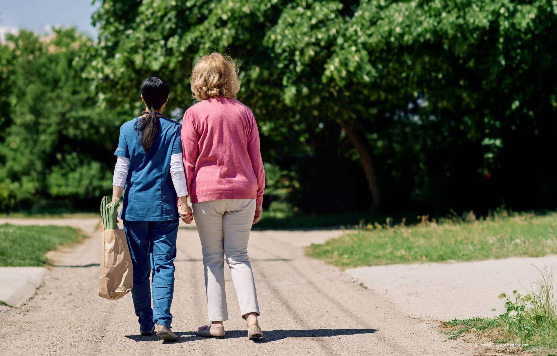 Eine Frau in einem rosa Pullover und ihre Betreuerin mit Einkäufen gehen Händchen haltend einen Weg entlang.
