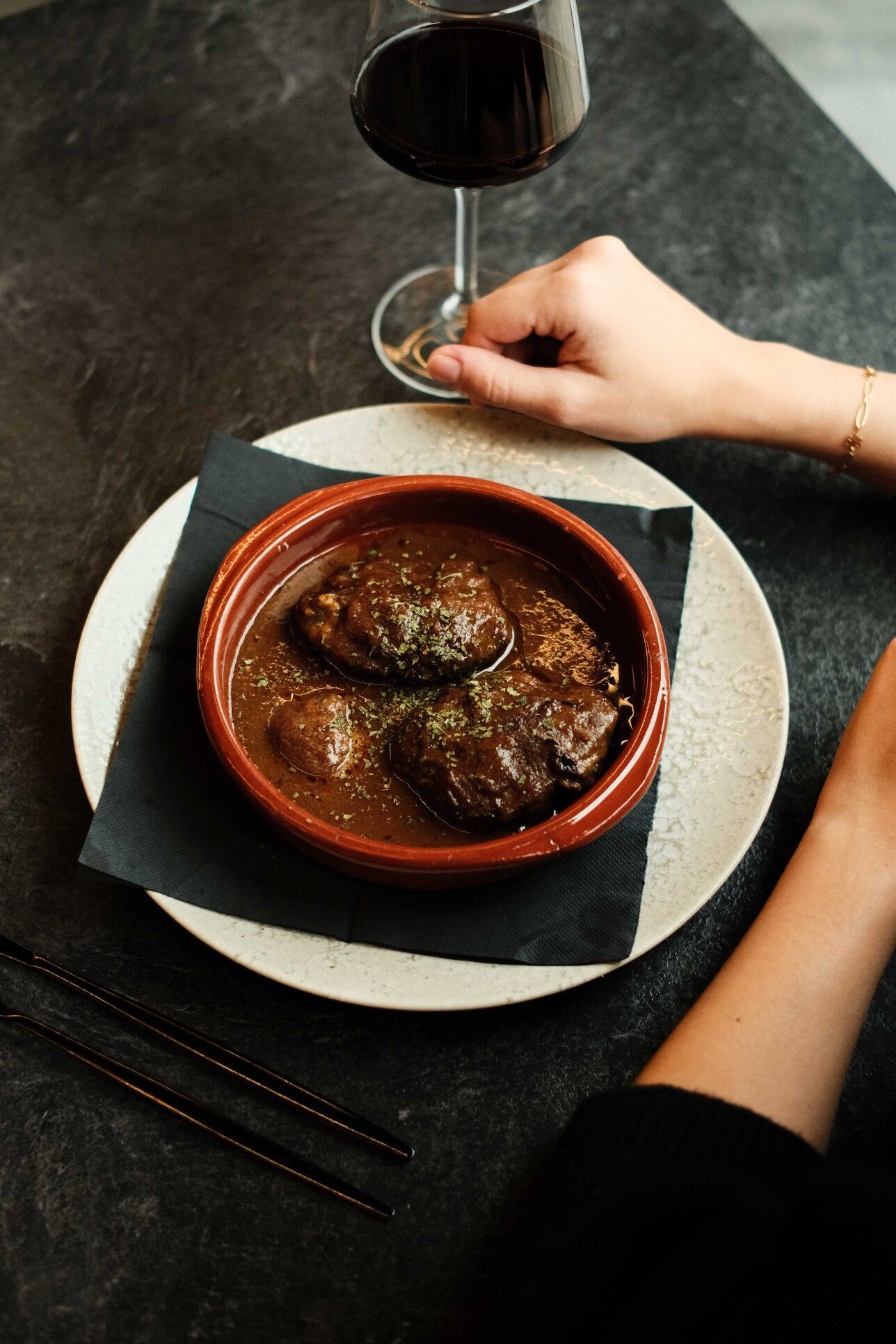 Un plato de estofado con una copa de vino tinto sobre una mesa. Una mano sostiene la copa.