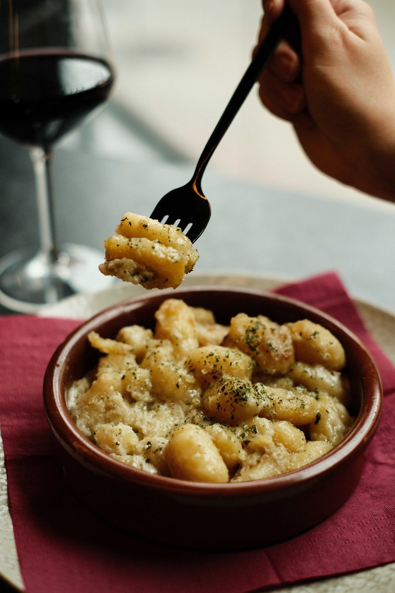 Ñoquis en un bol, con un tenedor en la mano. Vino tinto y una servilleta roja sobre la mesa.