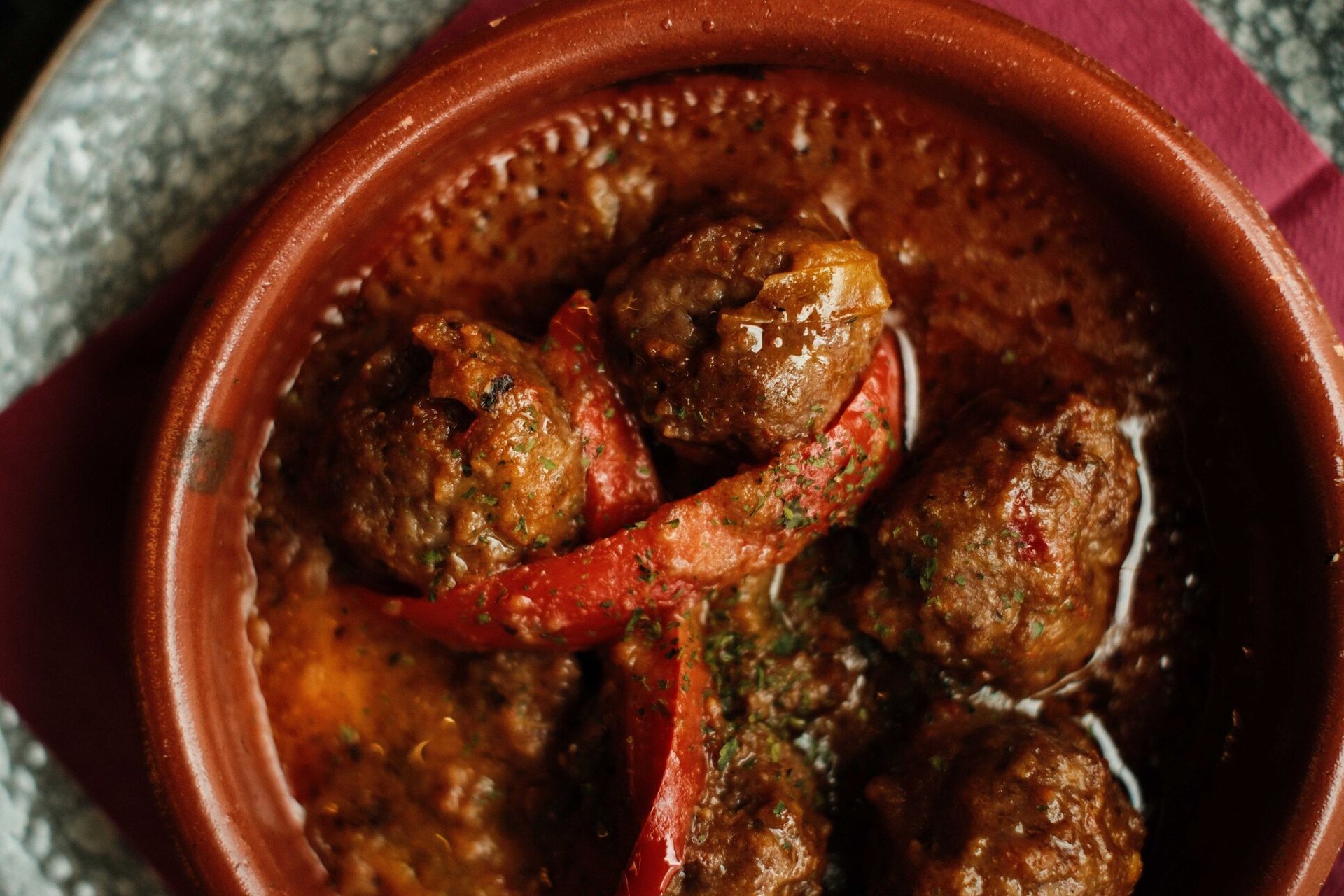 Albóndigas en salsa rica y marrón, servidas en un recipiente de terracota con rodajas de pimiento rojo.