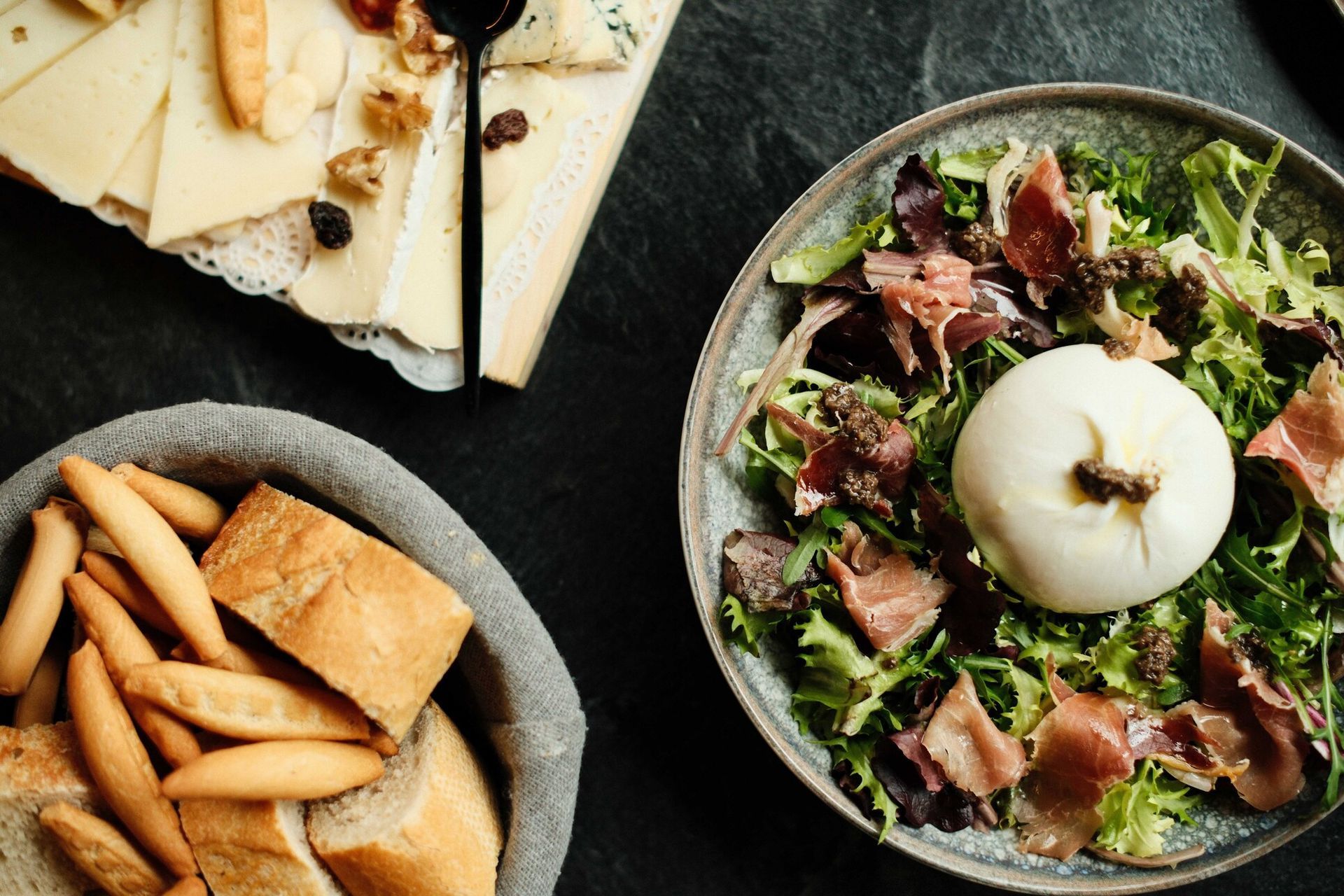 Tabla de quesos, cesta de pan y ensalada con burrata y prosciutto sobre una superficie oscura.