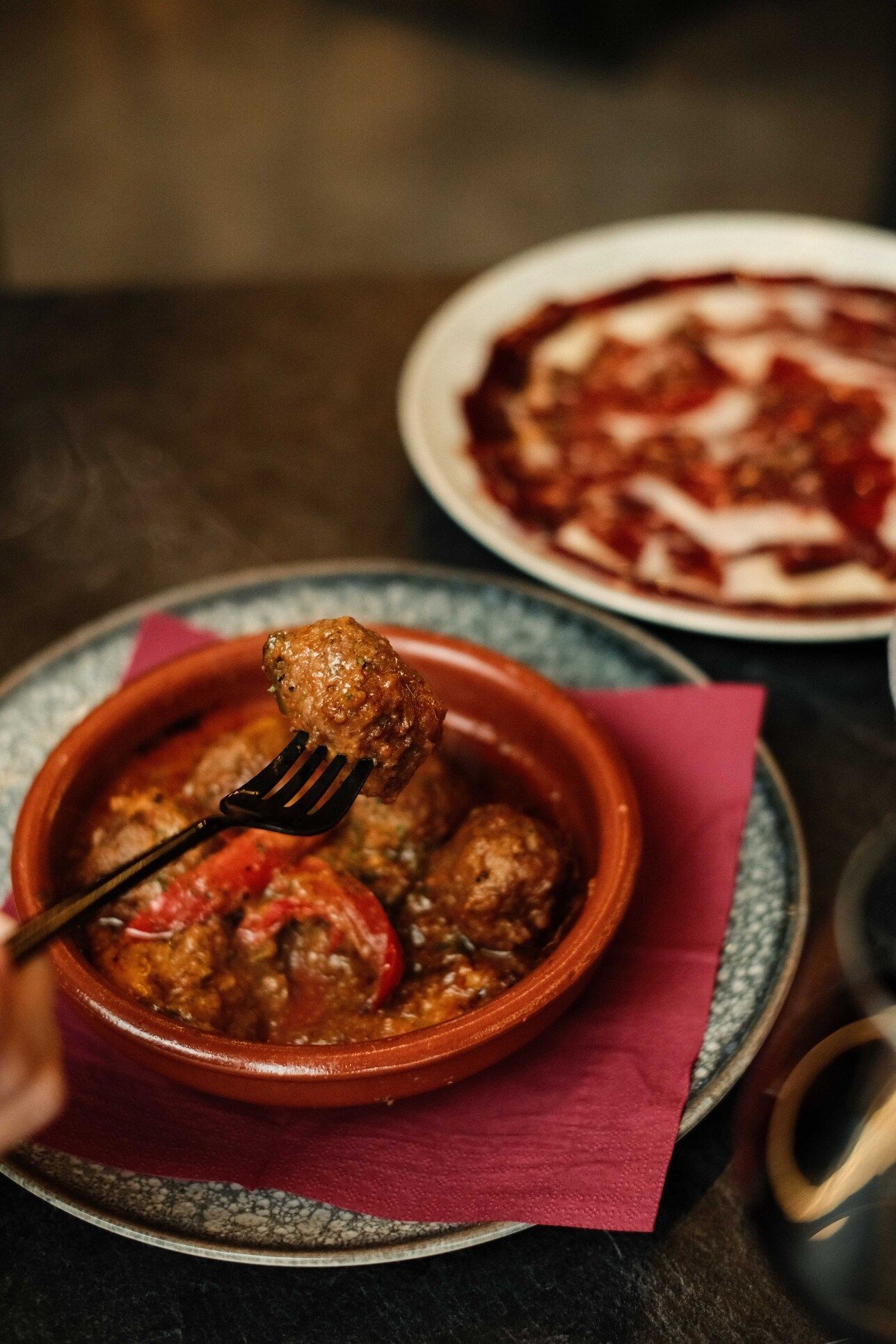 Albóndigas en un cuenco de terracota, levantadas con un tenedor. Detrás, un plato de embutidos.
