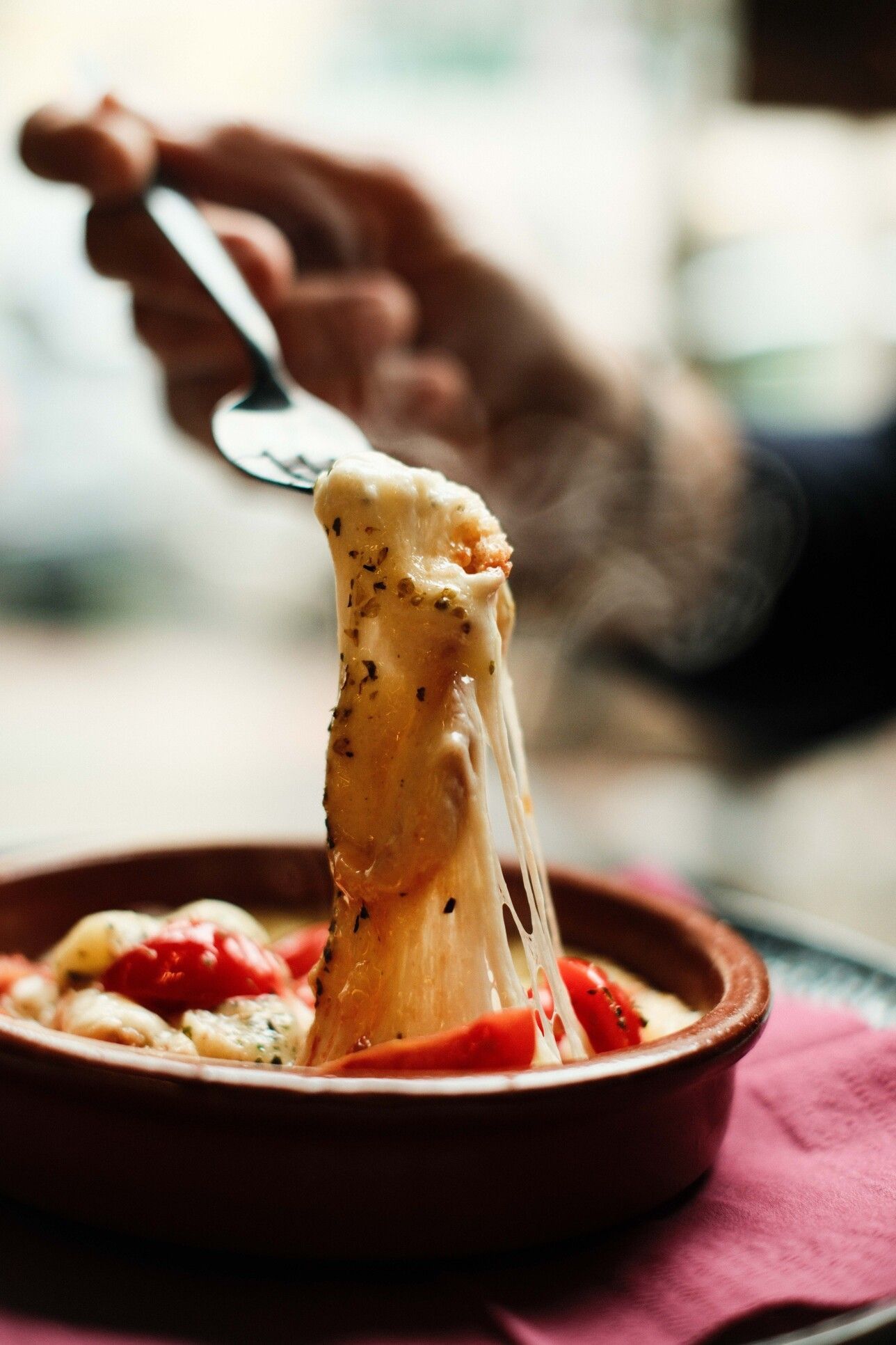 Plato de queso fundido con tomates, sacado con una cuchara de un pequeño tazón marrón. Saliendo vapor.