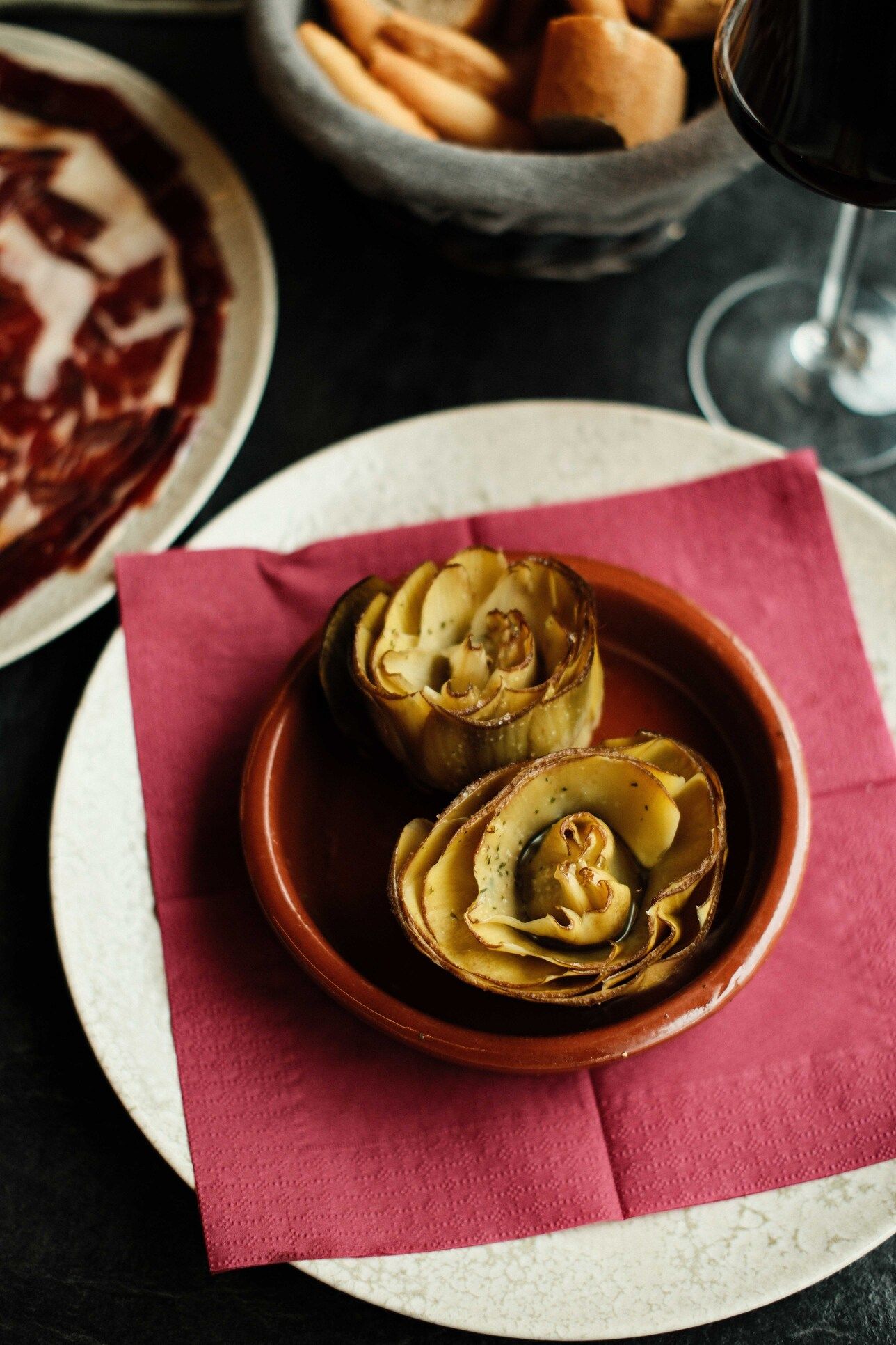 Corazones de alcachofa en un plato pequeño marrón sobre una servilleta roja, servidos con otras tapas y vino.