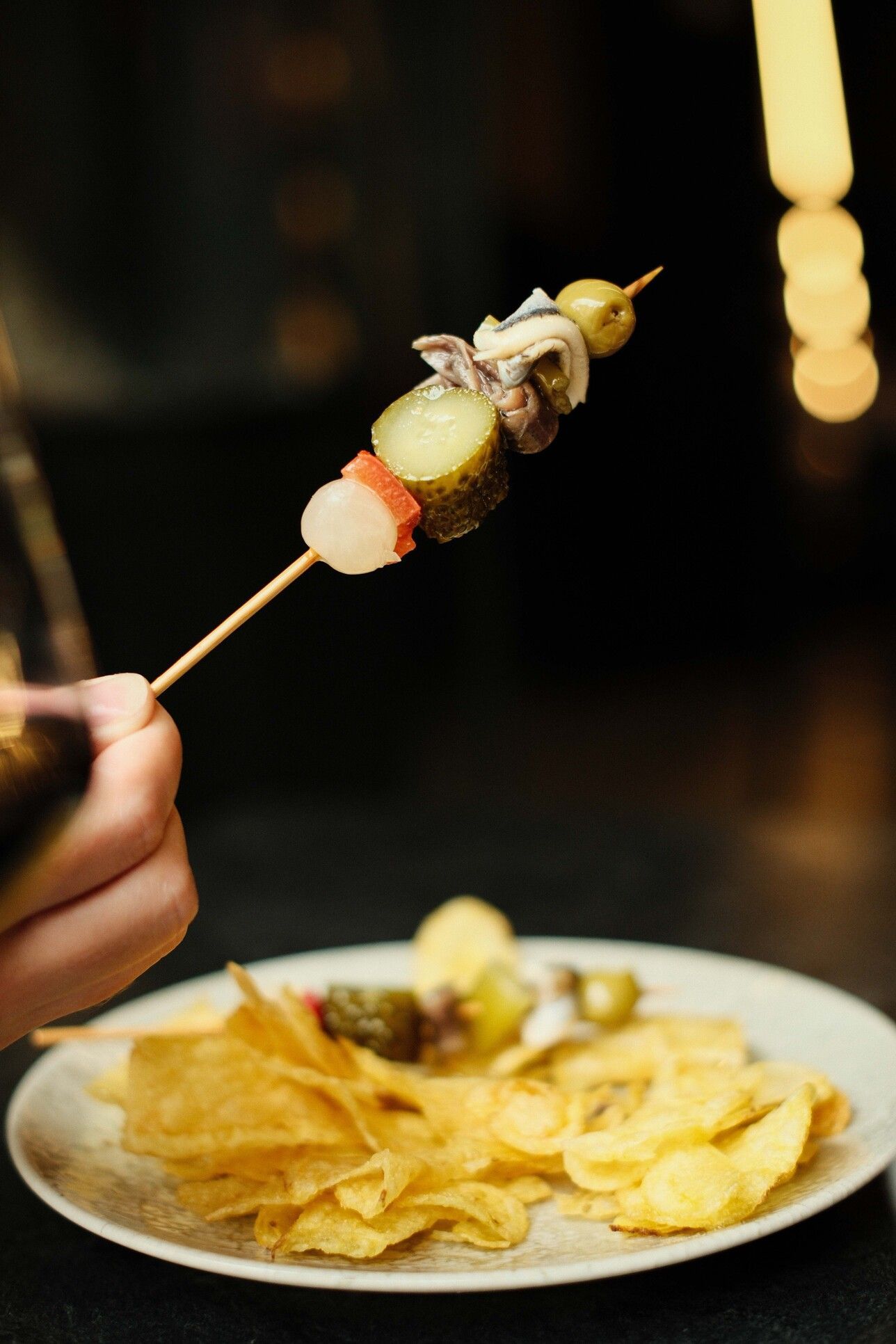 Mano sosteniendo una brocheta con encurtidos (aceitunas, pepinillos, anchoas) y patatas fritas en un plato.