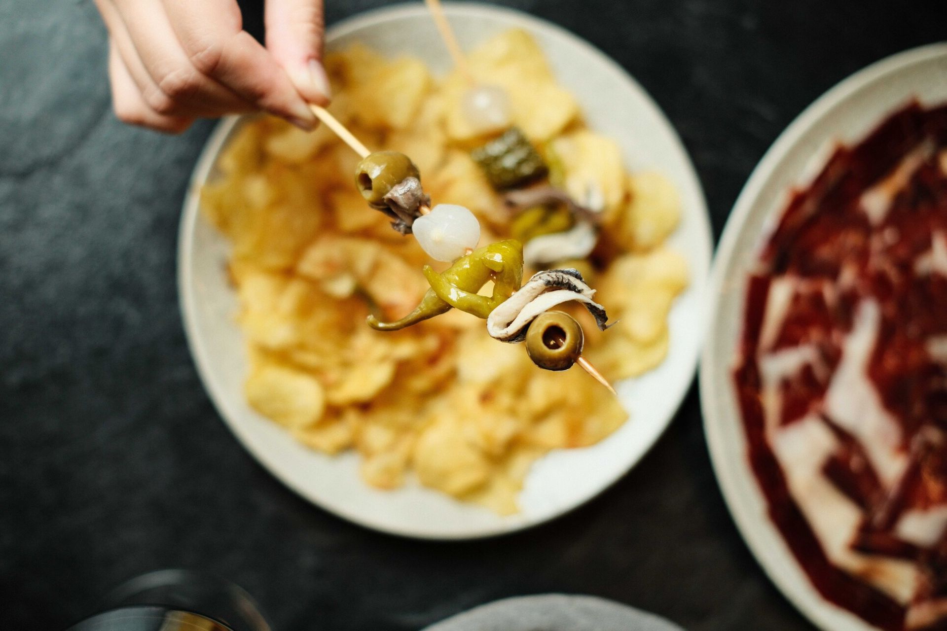 Mano sosteniendo una brocheta con aceitunas, pimientos y calamares sobre un plato de patatas fritas, con un plato de carne en rodajas cerca.
