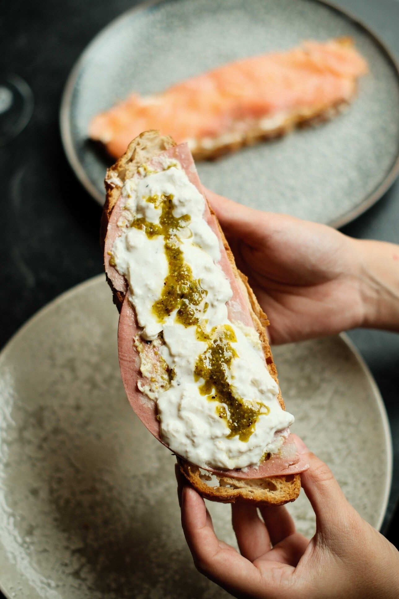 Manos sosteniendo un sándwich abierto con ricotta, jamón y pesto. En el segundo plato, salmón empanizado.