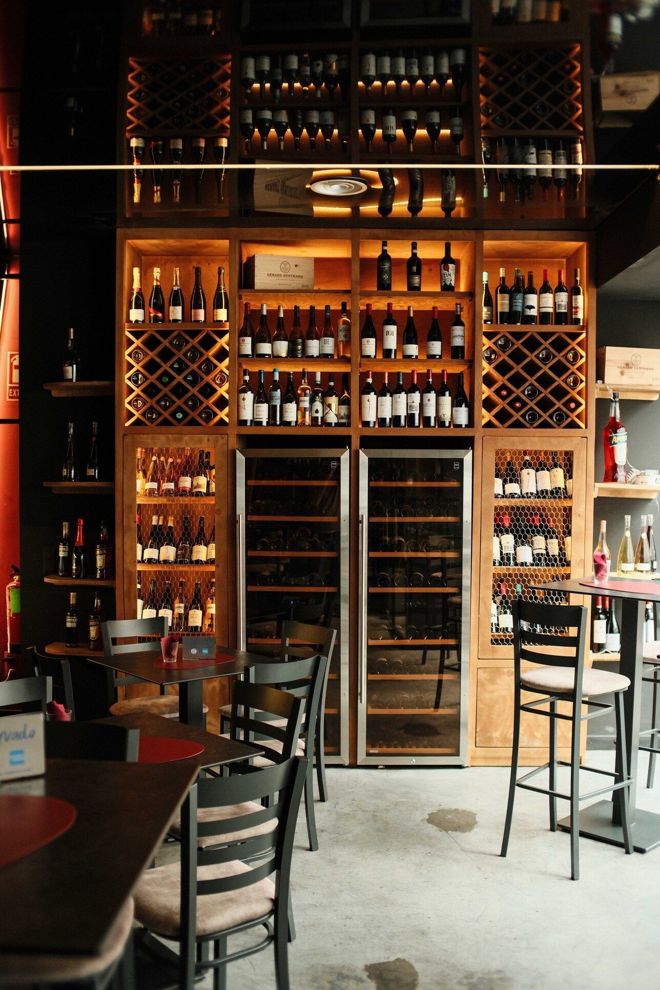 Botellas de vino expuestas en estanterías de madera y vitrinas refrigeradas en un bar de vinos. Mesas y sillas al frente.