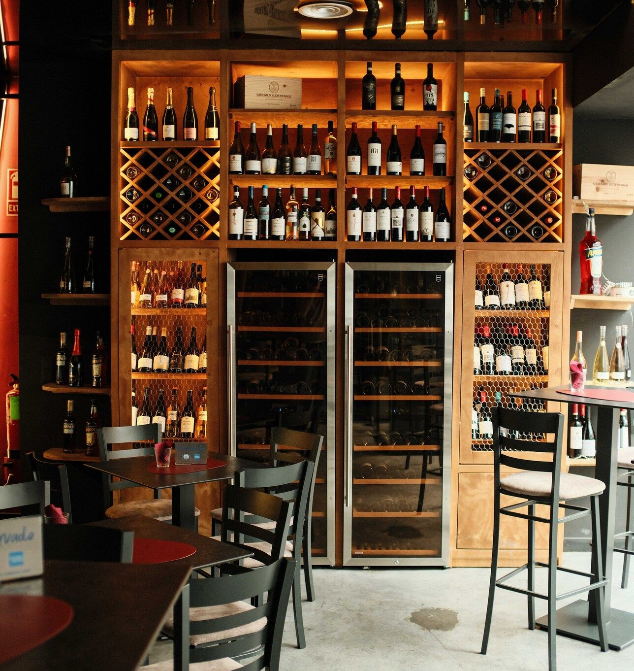 Interior del bar de vinos: estantes de madera llenos de botellas de vino, dos enfriadores de vino y mesas con sillas.