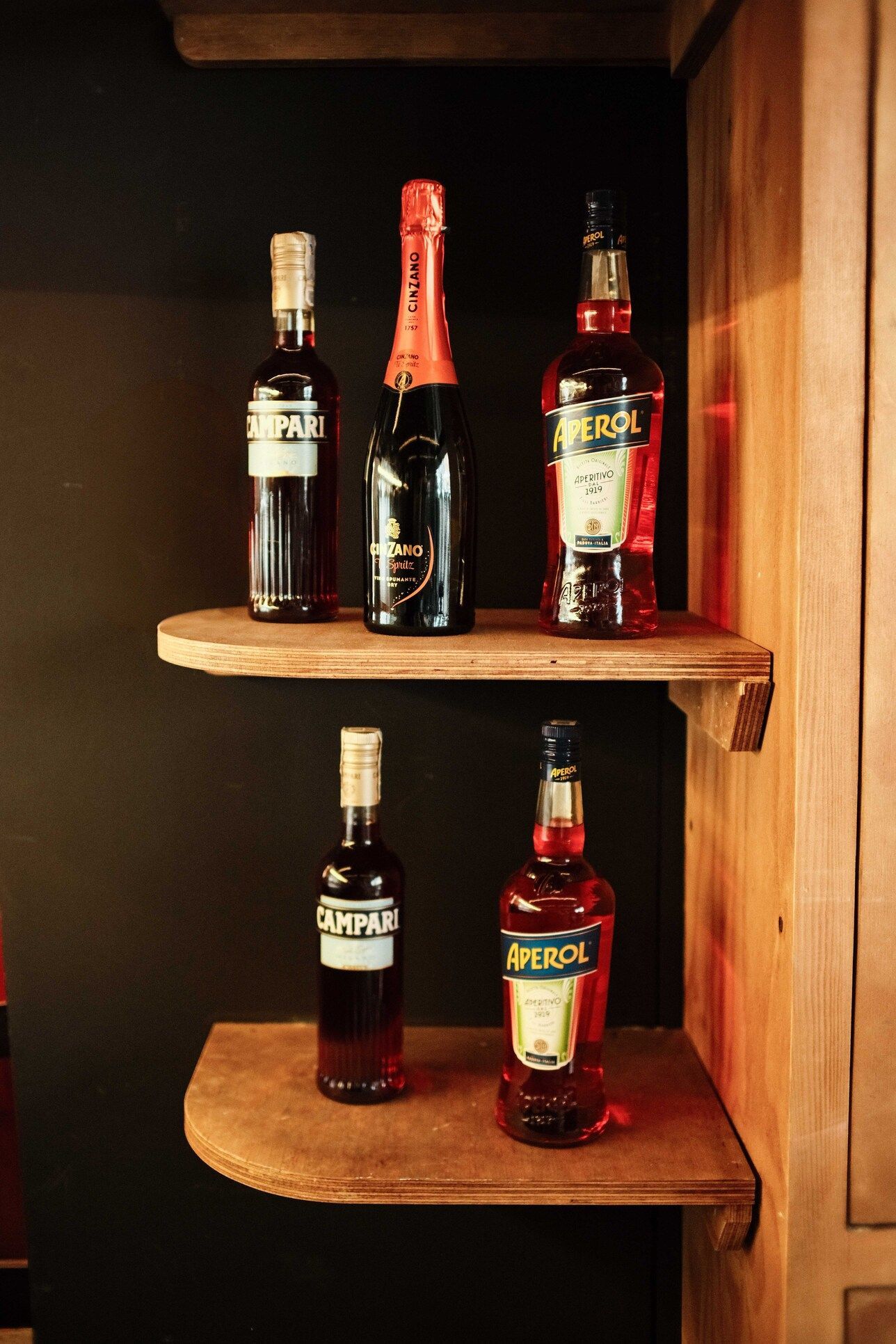 Botellas de licor en dos estantes de madera, contra una pared oscura. Incluye Aperol, Campari y vino espumoso.