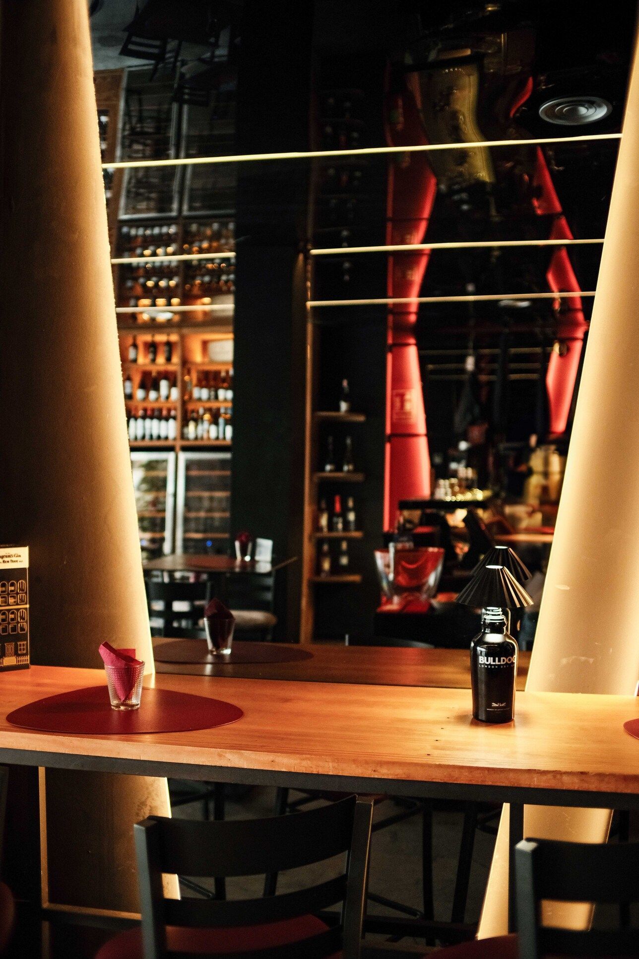 Interior de bar con mesa de madera, espejo reflectante y estantes con bebidas alcohólicas. Iluminación cálida.