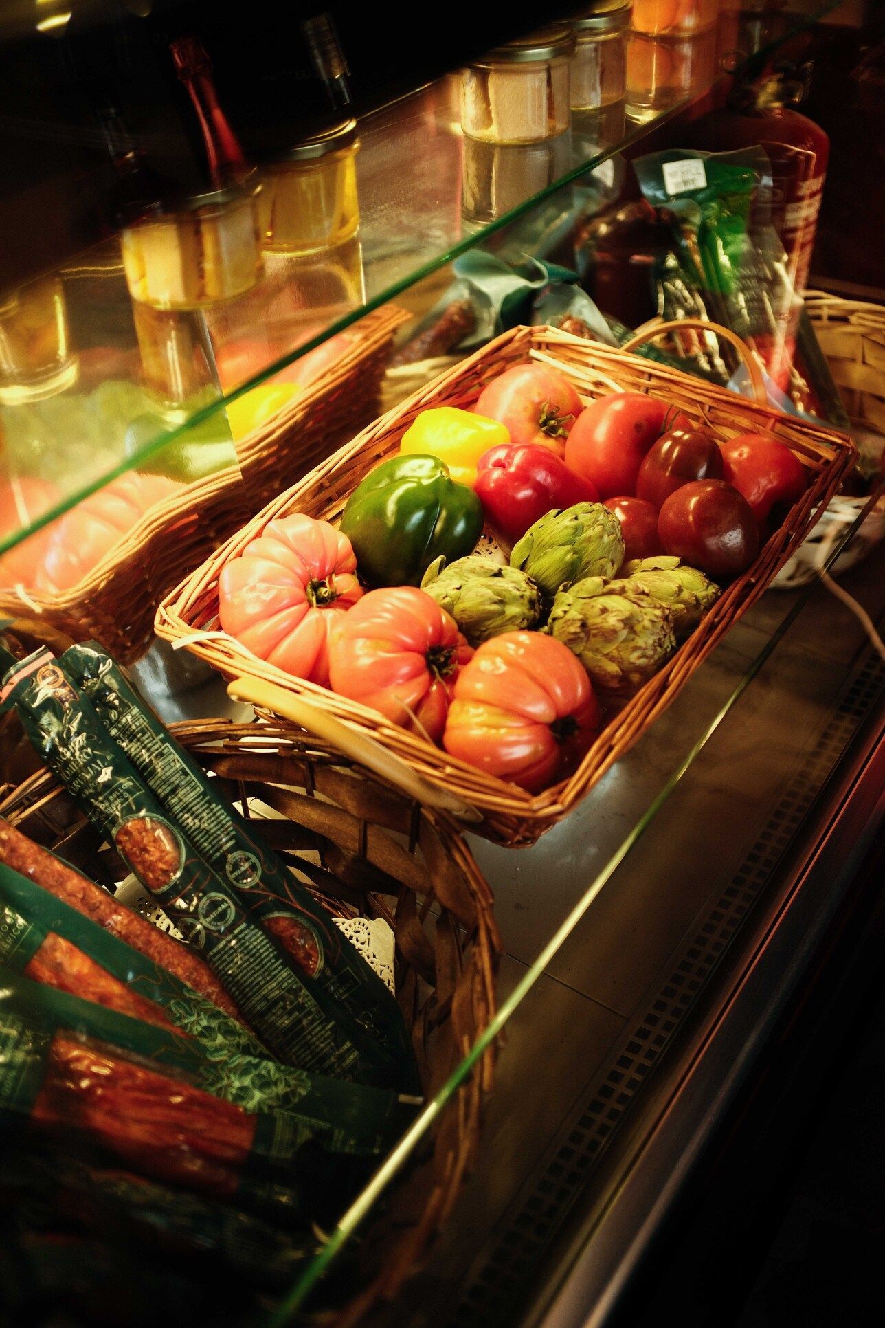 Vitrina con cestas de tomates, alcachofas y pimientos de colores. También hay paquetes de salchichas.