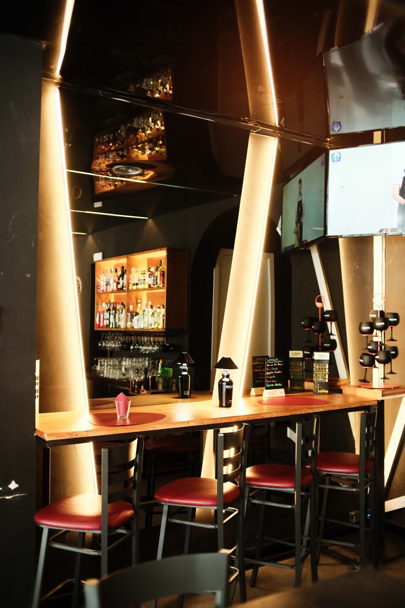 Interior de bar con barra de madera, taburetes rojos, vigas de soporte iluminadas y botellas en estantes.