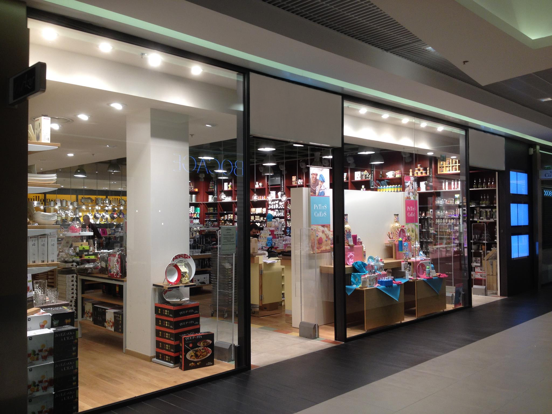 La façade d'un magasin de détail, avec ses parois vitrées, présente divers articles ménagers, des étagères et des produits dans un couloir de centre commercial.