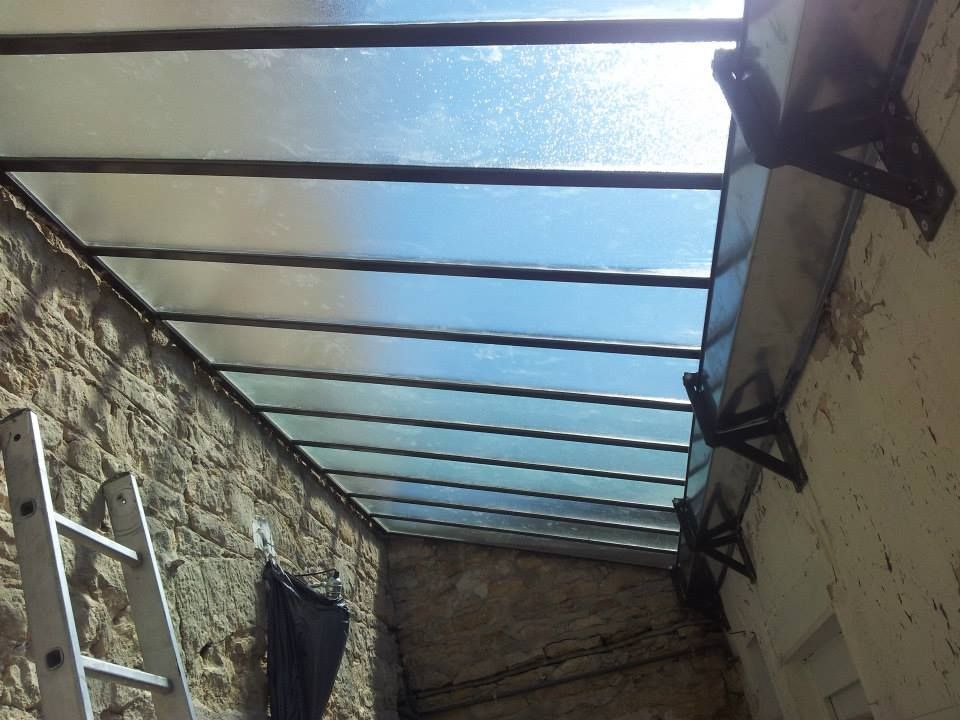 Un toit en appentis vitré, soutenu par des supports et une charpente en métal sombre, est installé entre deux murs de pierre, avec une échelle à proximité.