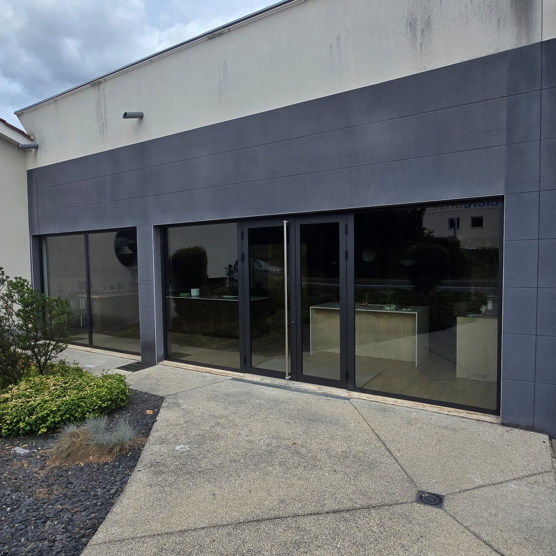 Vue extérieure de l'entrée d'un bâtiment avec des portes et fenêtres en verre teinté foncé, une façade grise et une allée pavée.