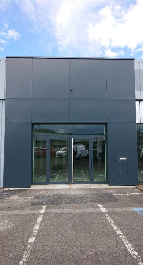 L'entrée d'un immeuble commercial avec des portes doubles vitrées intégrées à une façade gris foncé, vue depuis un parking asphalté.