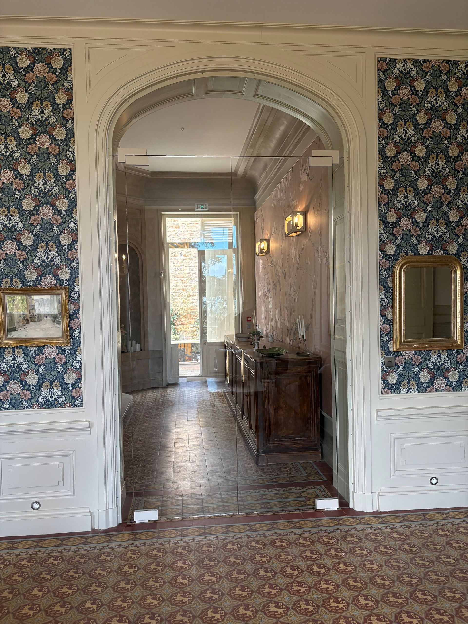 Couloir orné de papier peint à motifs floraux, porte cintrée, moquette à motifs et meubles en bois au-delà