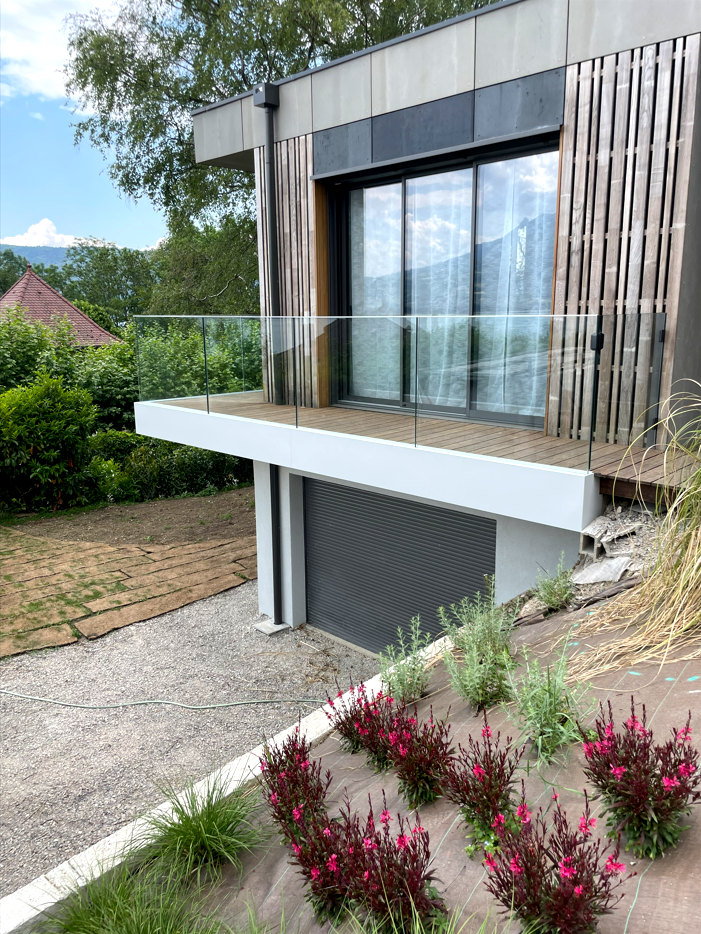 Maison moderne avec balcon, balustrade en verre et grandes portes coulissantes en verre, offrant une vue sur le jardin et les montagnes.