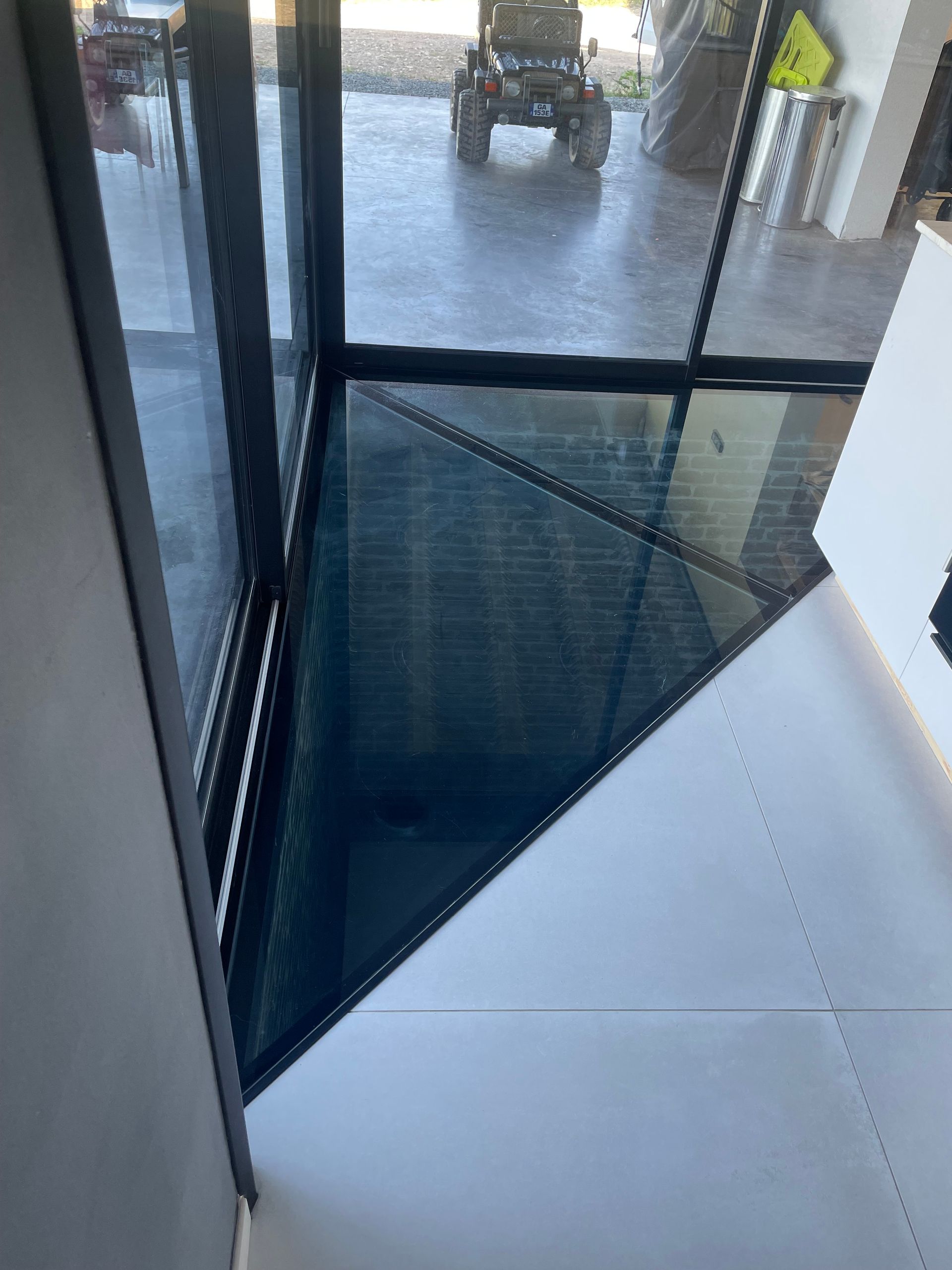 Un panneau de sol triangulaire en verre encastré dans un carrelage blanc, près d'une grande fenêtre donnant sur un espace sombre.